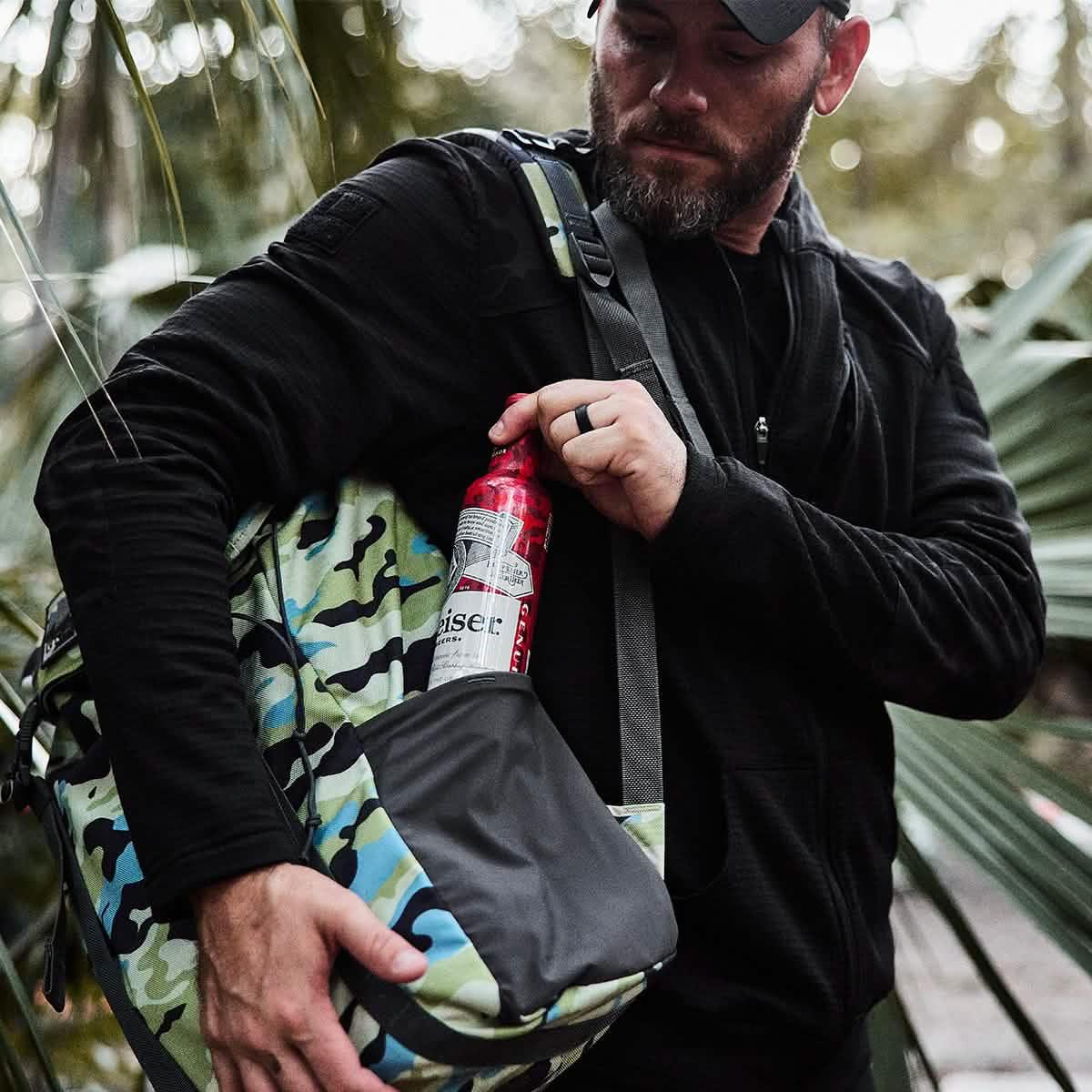 Man in black jacket placing a red bottle into side pocket of Everglades camo GORUCK rucking backpack outdoors