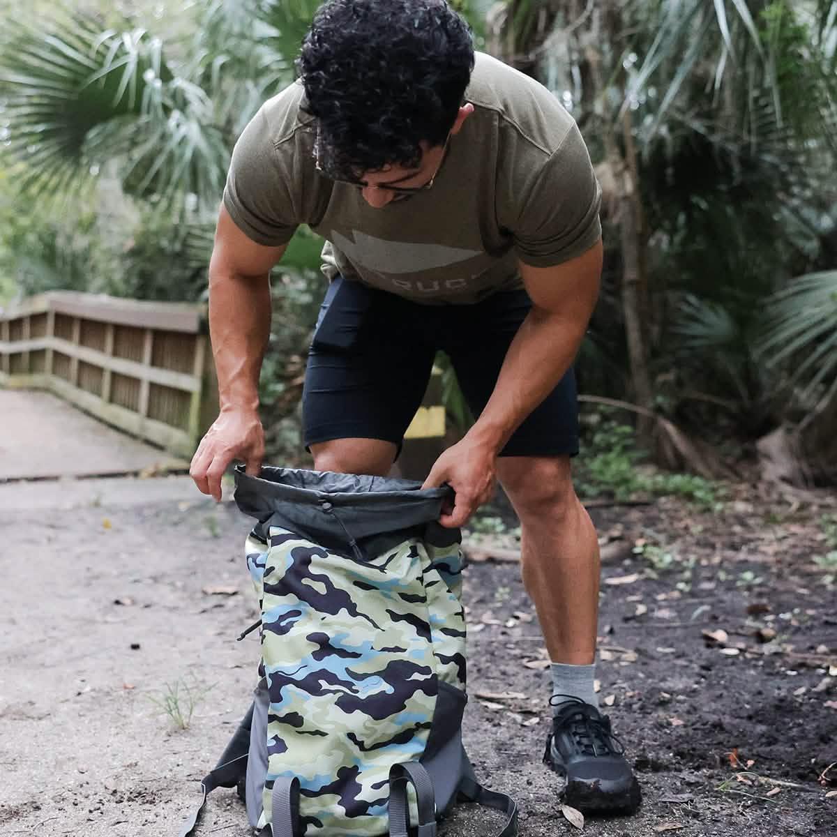 Man outdoors packing camo GORUCK backpack on forest trail near wooden bridge