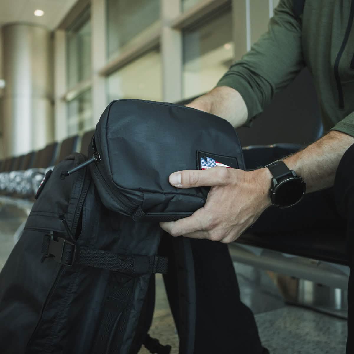 In an airport terminal, a traveler sits holding a sleek black travel bag adorned with a distinctive American flag patch. They are dressed in a green shirt and wear a black smartwatch that perfectly complements their GORUCK GR1 Field Pocket - Dyneema electronics organizer tucked inside.