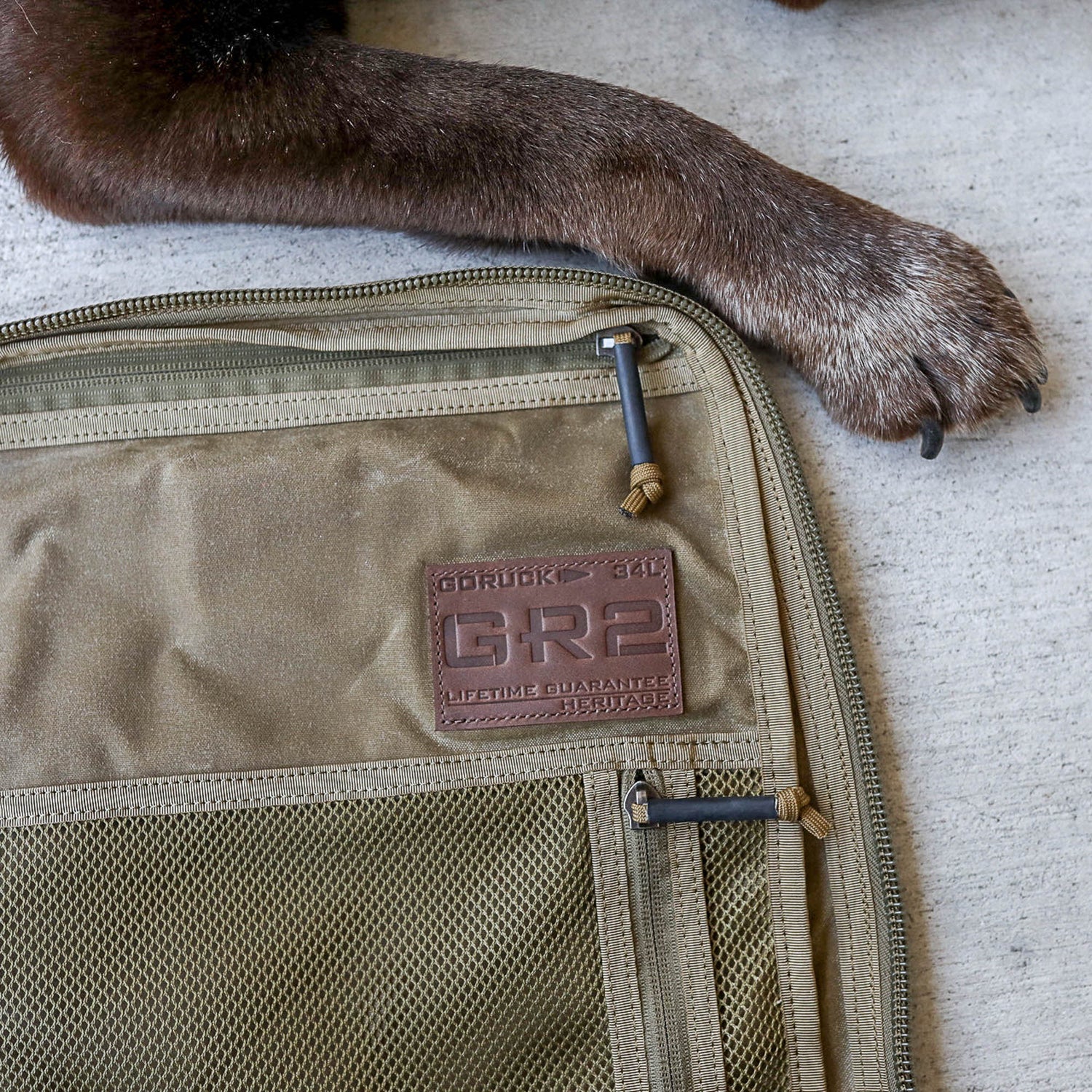 A dog's paw rests beside a tan GR2 - Heritage Waxed Canvas backpack with a Martexin wax finish, set on a concrete surface.
