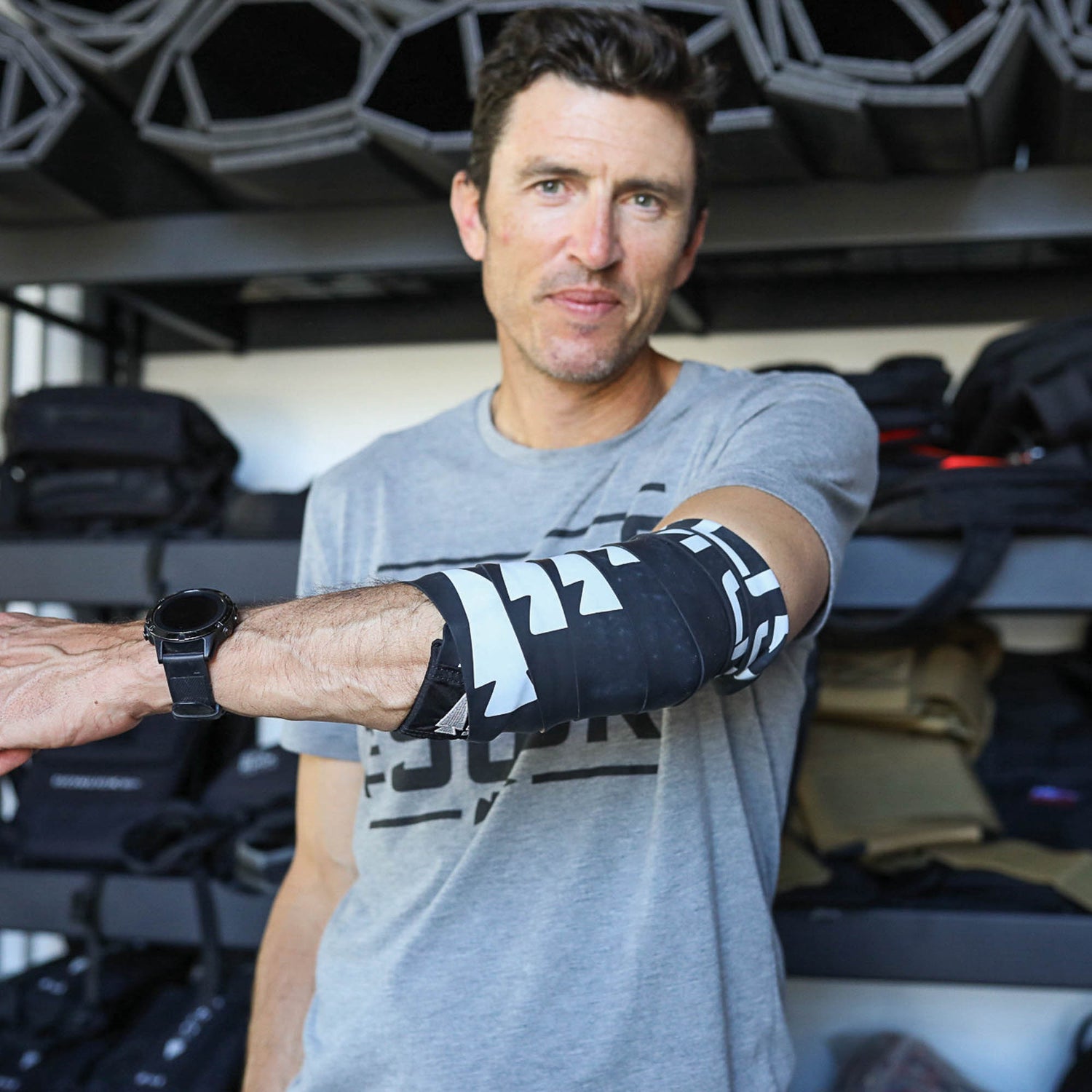 A man in a gray t-shirt displays the Recovery Floss Band, a black and white arm brace that uses muscle compression to improve mobility, while standing before shelves lined with bags.