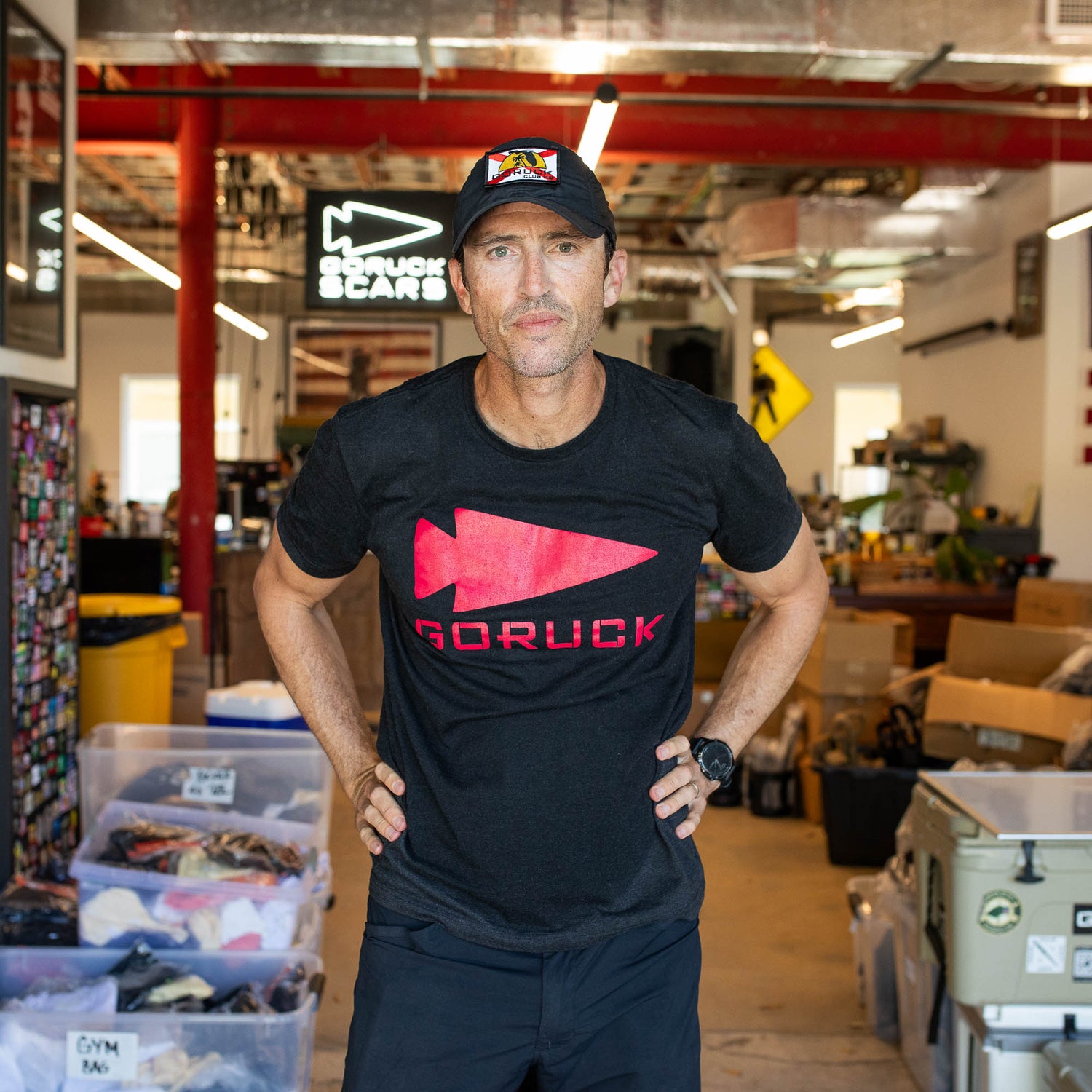 A man wears the GORUCK Spearhead Tee - Tri-Blend and a cap, standing in a warehouse with hands on hips, channeling Special Forces confidence as he looks at the camera.
