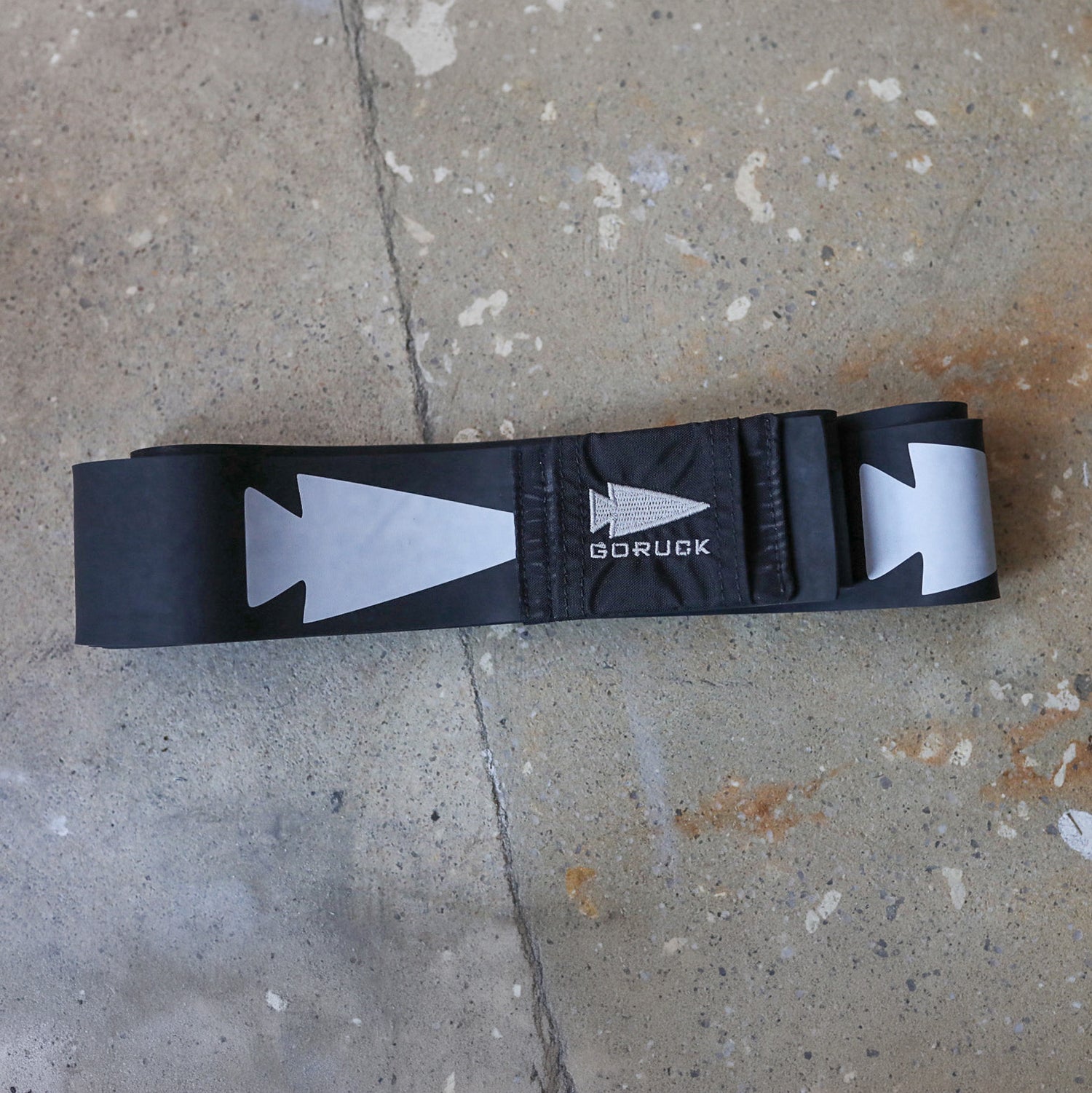 A black Recovery Floss Band with reflective white arrows, displayed on a concrete floor, ideal for pairing with compression gear to boost mobility and aid muscle recovery during active days.