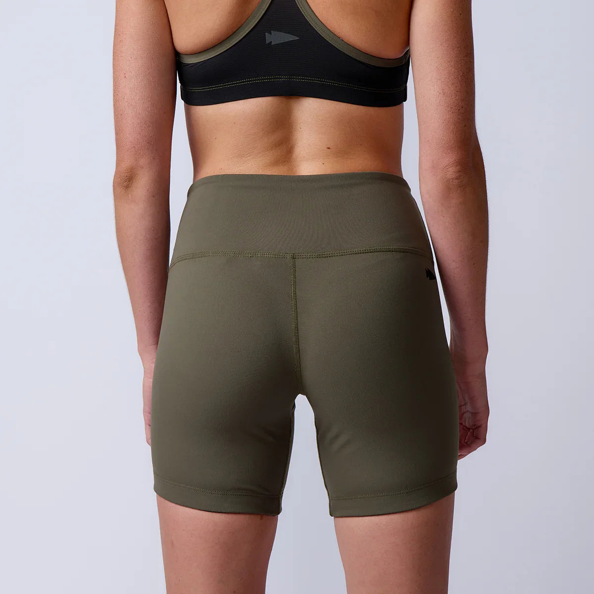 Back view of a woman wearing olive green high-waisted sports shorts and a black sports bra against a plain background