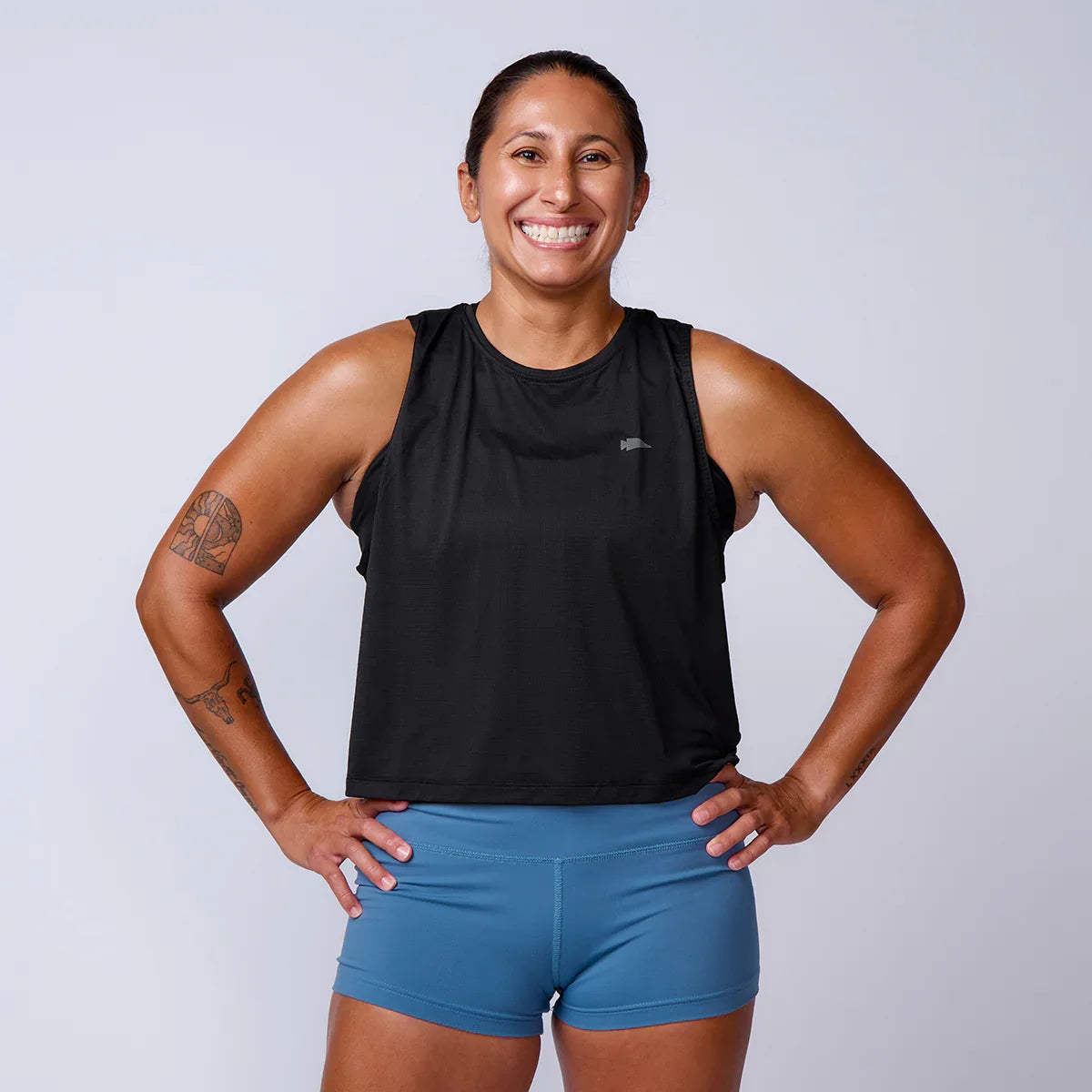 Smiling woman in athletic GORUCK gear, black tank top and blue shorts, studio background