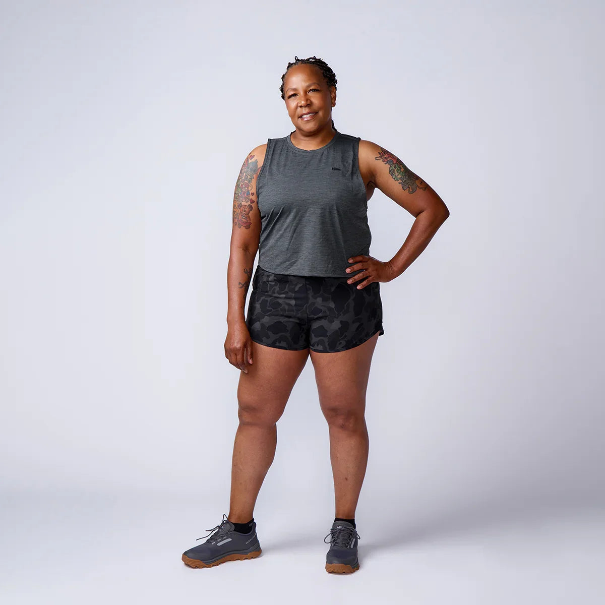 Woman in gray sleeveless top, black camo shorts, and gray GORUCK shoes posing on white background
