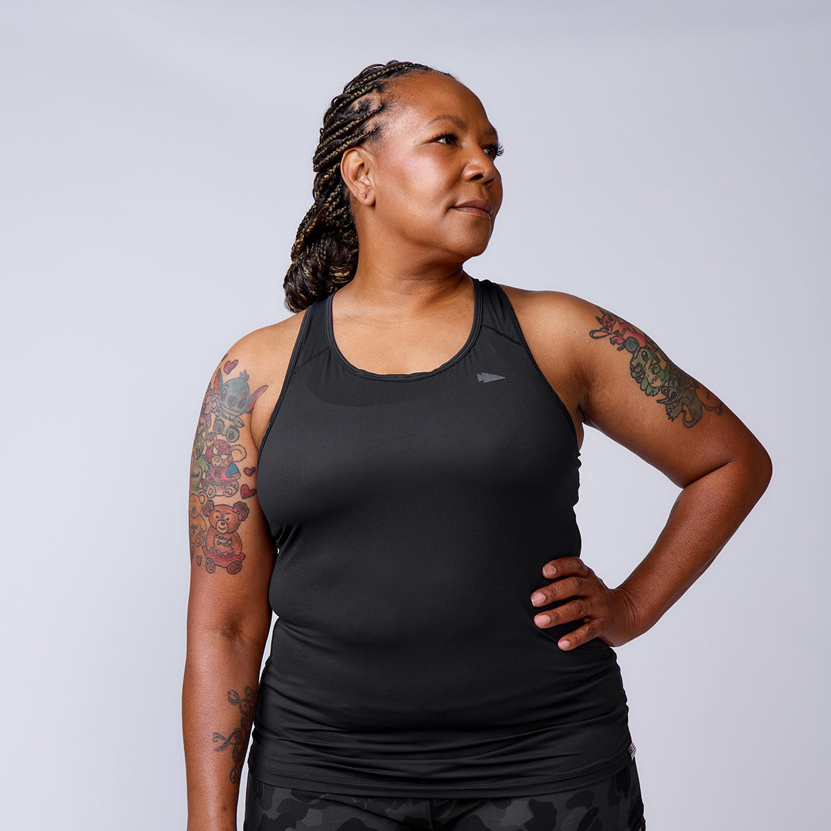 A woman with braided hair and tattoos poses confidently in the Women’s Racerback Tank - ToughMesh, set against a plain background.