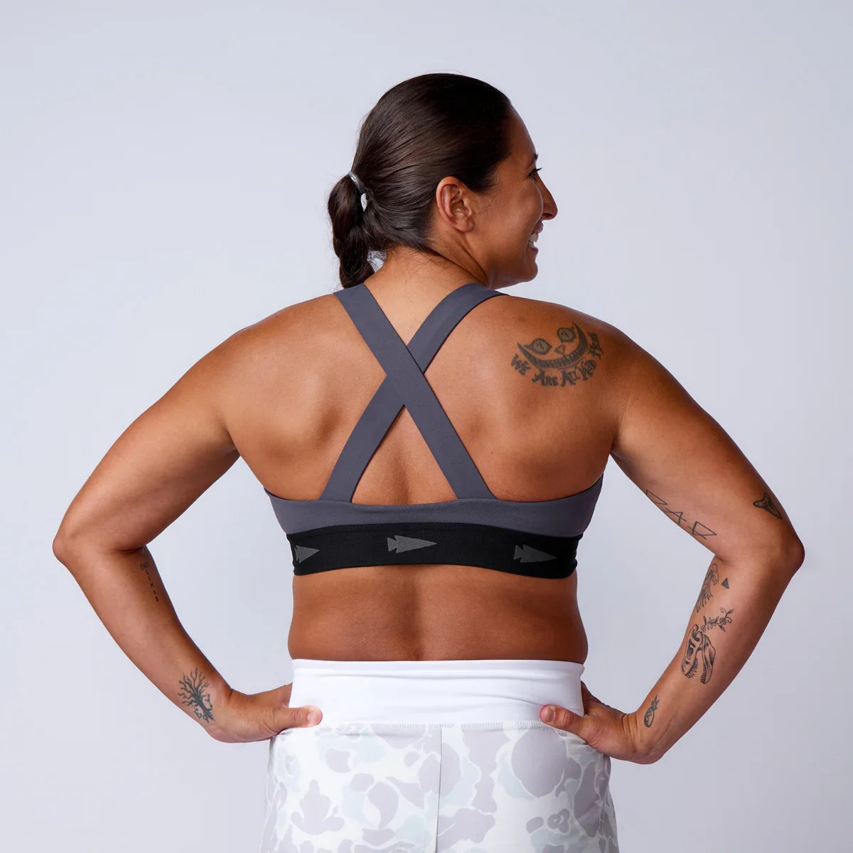 Woman wearing gray and black cross-back sports bra and white patterned leggings showing back tattoos