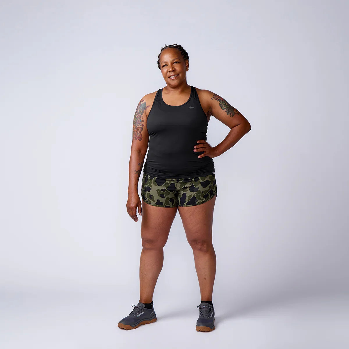 Woman in black tank top, camo shorts, and sneakers stands confidently against gray background with tattoos on arms