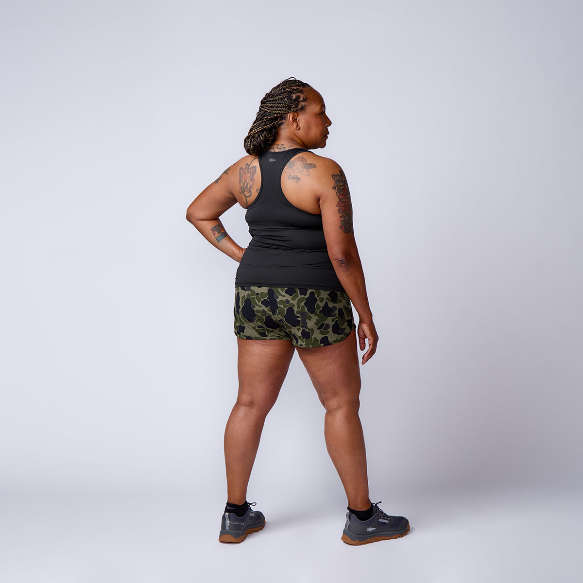 A person stands in a studio, facing away, wearing a black tank top, Women’s Training Shorts - ToughStretch in camo, and sneakers.