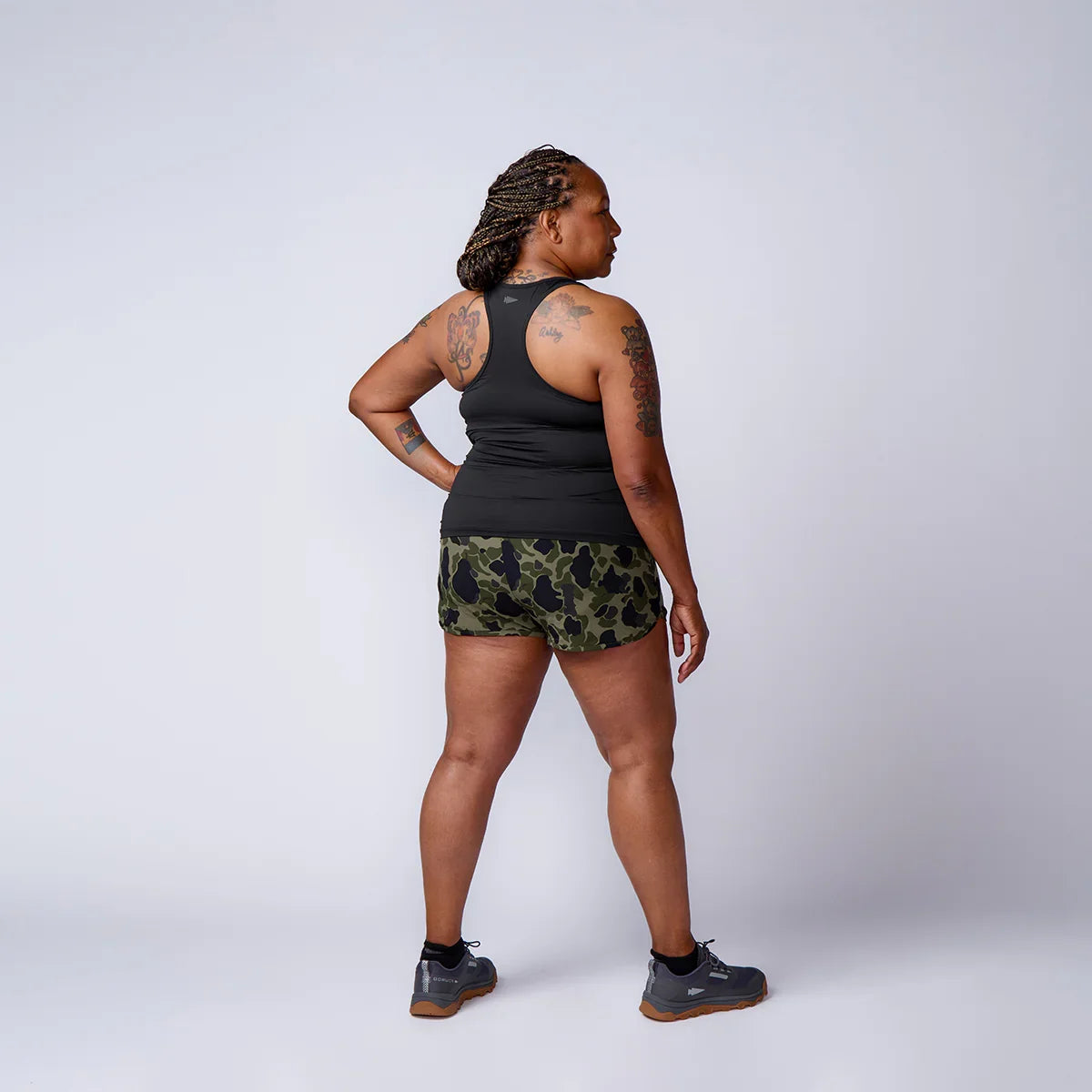 Woman with tattoos wearing black tank top, camo shorts, and gray GORUCK shoes standing against white background