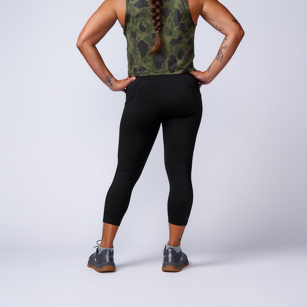 Person stands with hands on hips, wearing ToughFlex Women’s Training Leggings Pocket, a camo tank top, and gray sneakers, facing away.