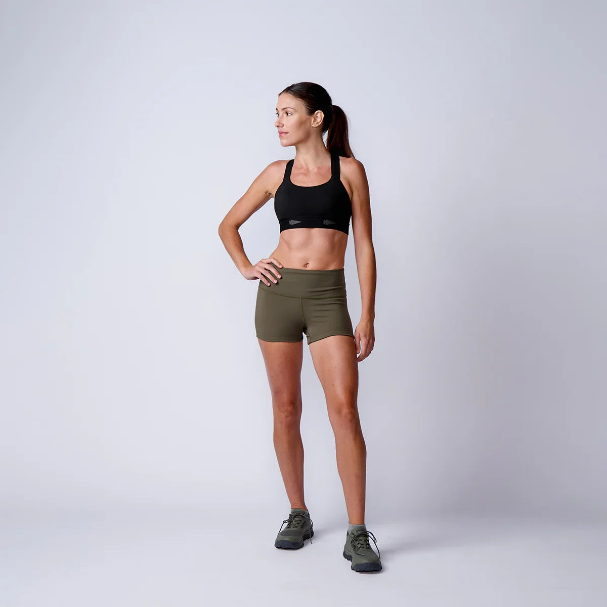 Fit woman standing in black sports bra, olive green shorts, and hiking shoes on plain background