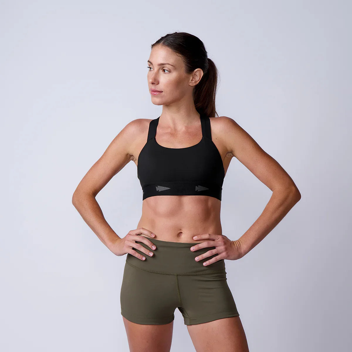 Fit woman wearing a black sports bra and olive green shorts standing against a plain light gray background
