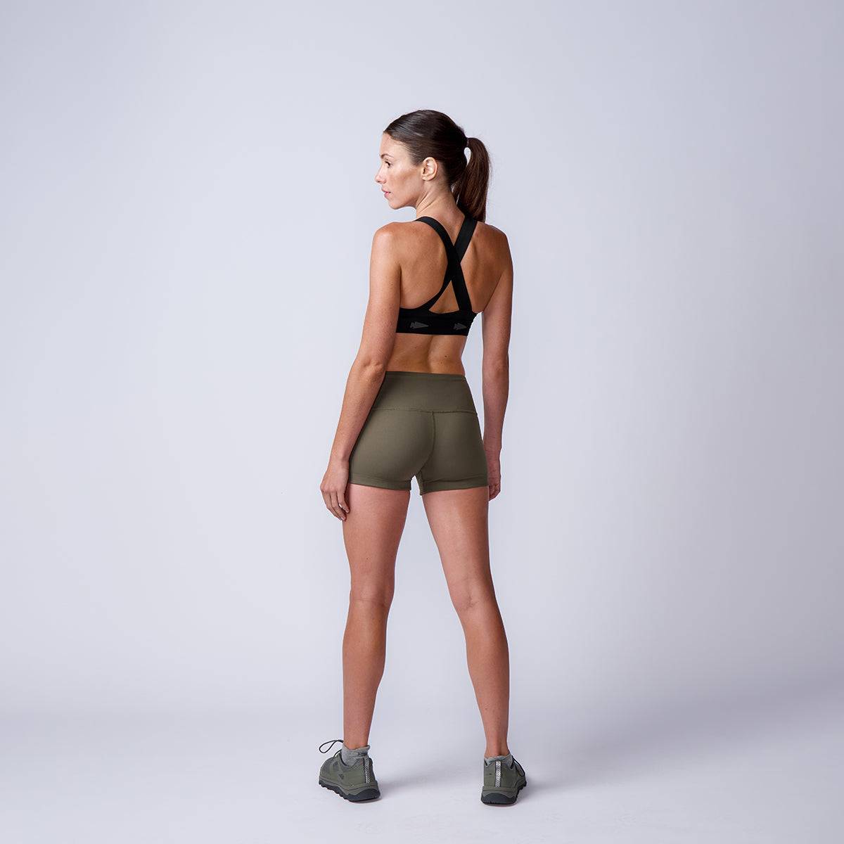 Woman in athletic wear stands facing away, wearing a black sports bra and high-waisted green Women’s Squat Shorts - ToughFlex.