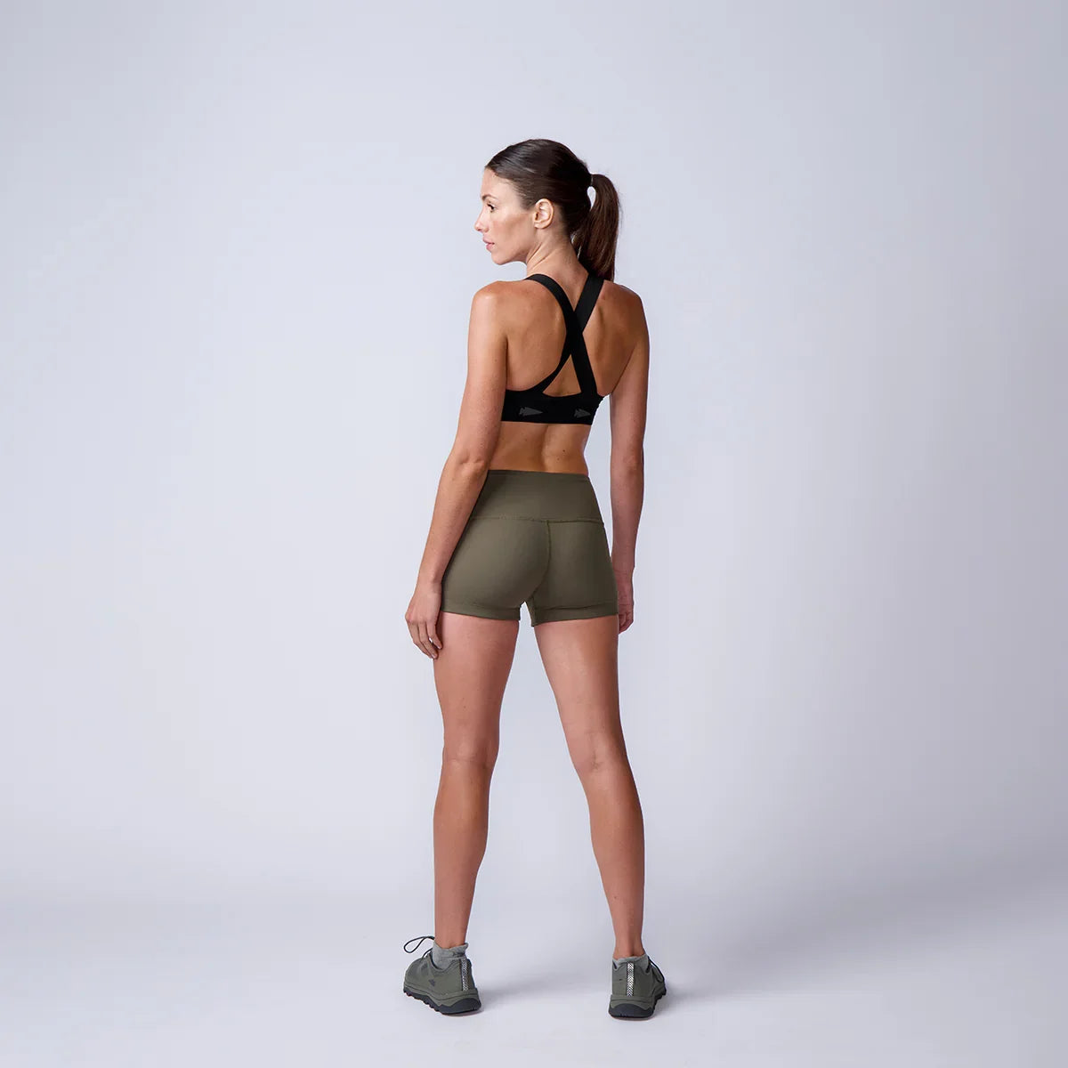 Back view of fit woman in black sports bra and olive rucking shorts with hiking shoes on plain background