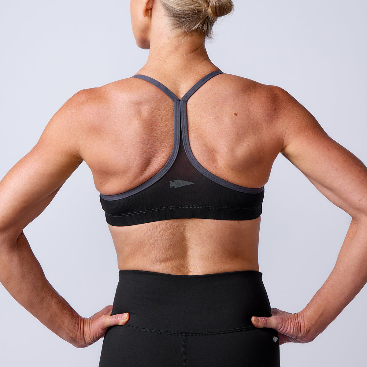 A woman is shown from behind wearing the Stealth Bra - ToughFlex, a minimalist sports bra with lightweight support, paired with high-waisted leggings and her hands on her hips.