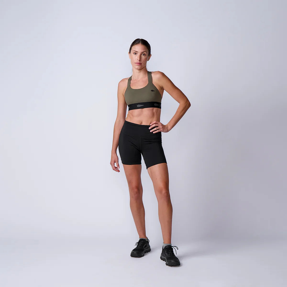 Fit woman in olive green sports bra and black workout shorts posing in studio setting with neutral background