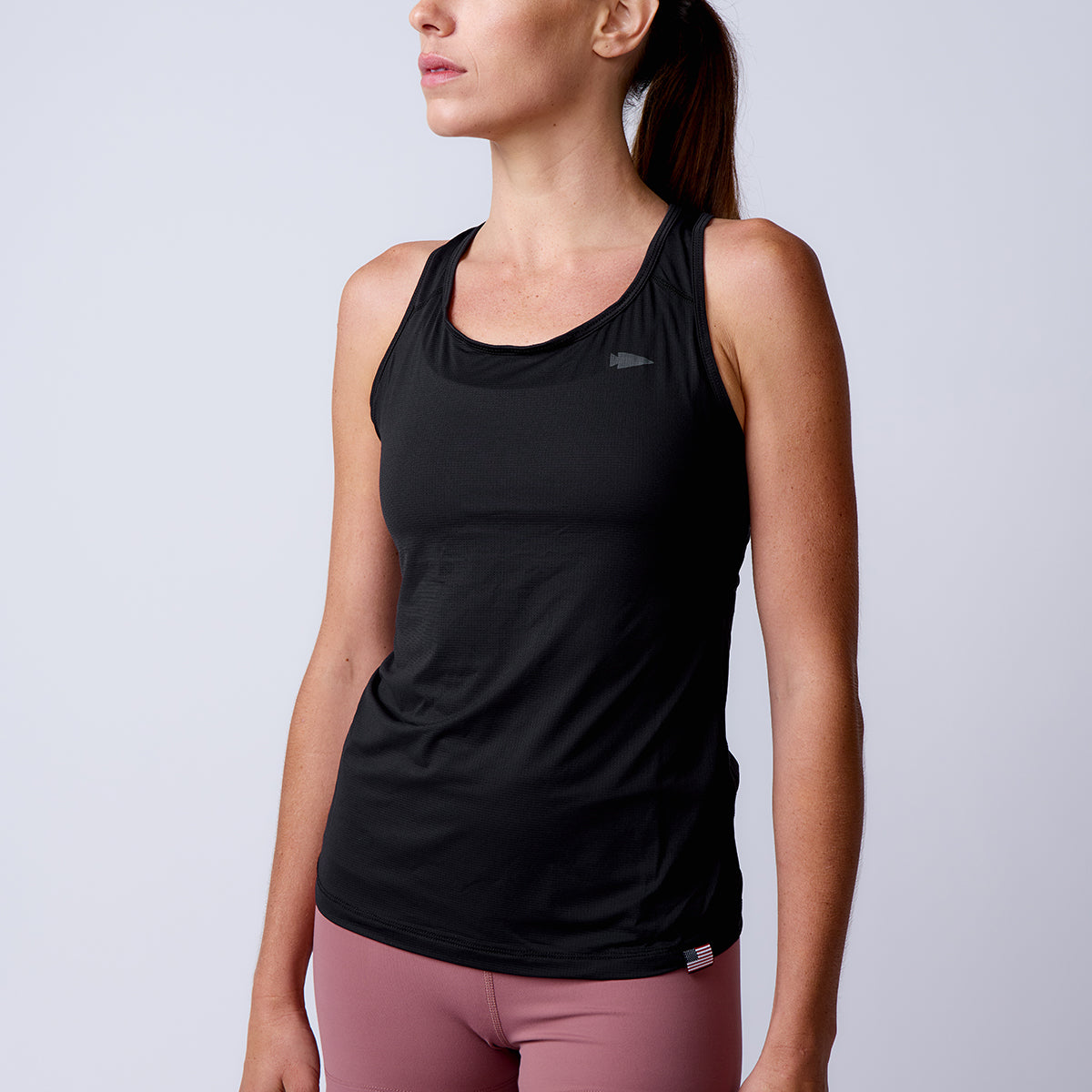 A woman wears the Women’s Racerback Tank - ToughMesh in black, crafted from Italian performance fabric, paired with pink leggings, standing against a plain light background.