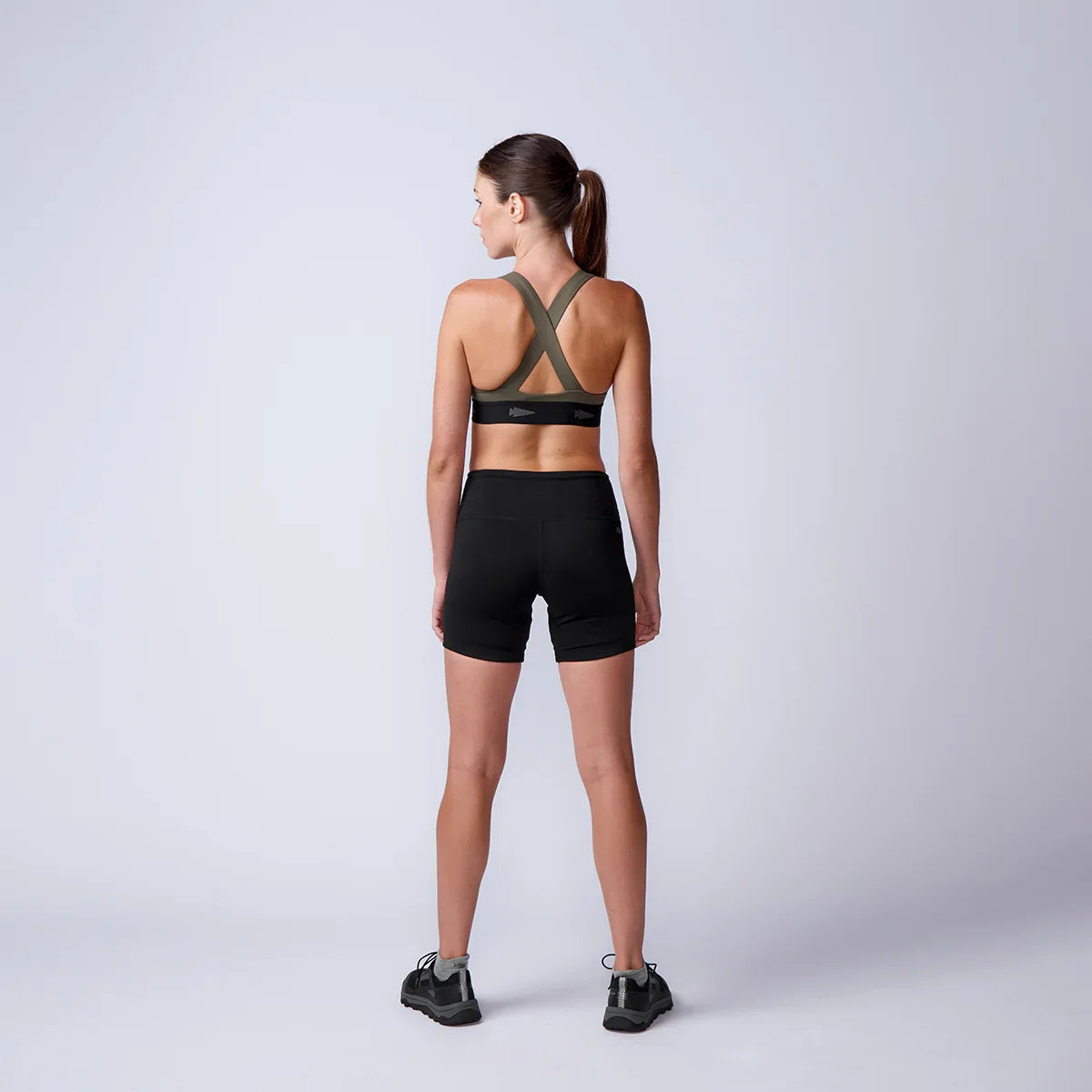 Athletic woman in black sports bra and shorts standing facing away on plain studio background for GORUCK fitness apparel