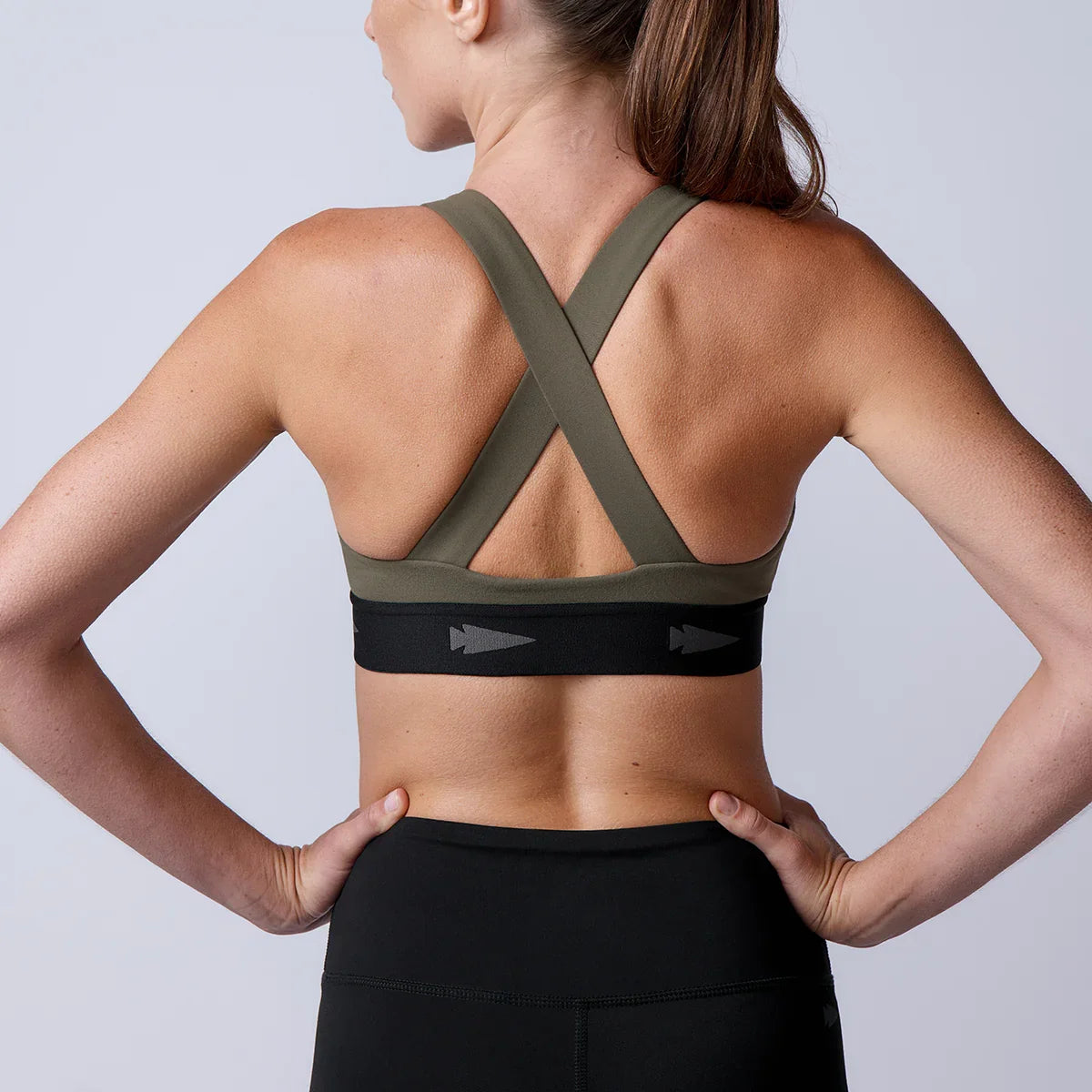 Back view of woman wearing olive green and black crisscross sports bra with black leggings against plain background