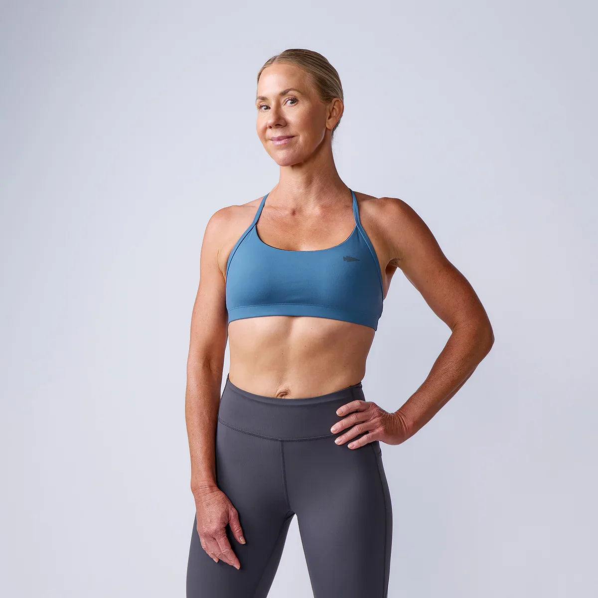 fit woman in blue sports bra and gray leggings standing confidently in studio against light background
