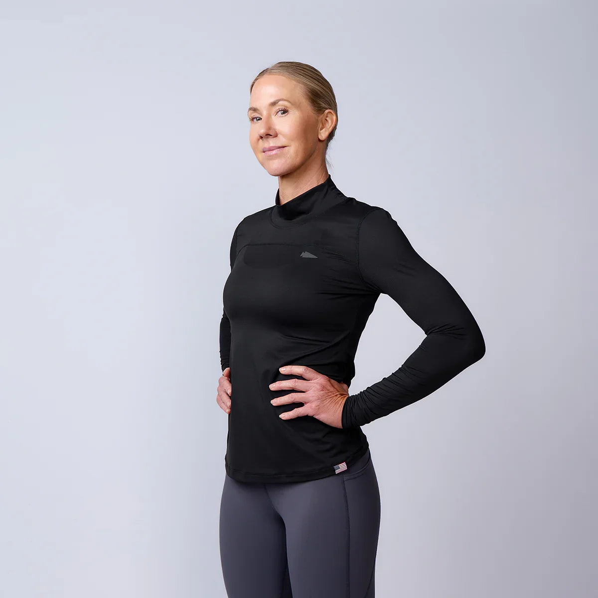 Woman wearing black long-sleeve athletic top and dark gray leggings posing with hands on hips