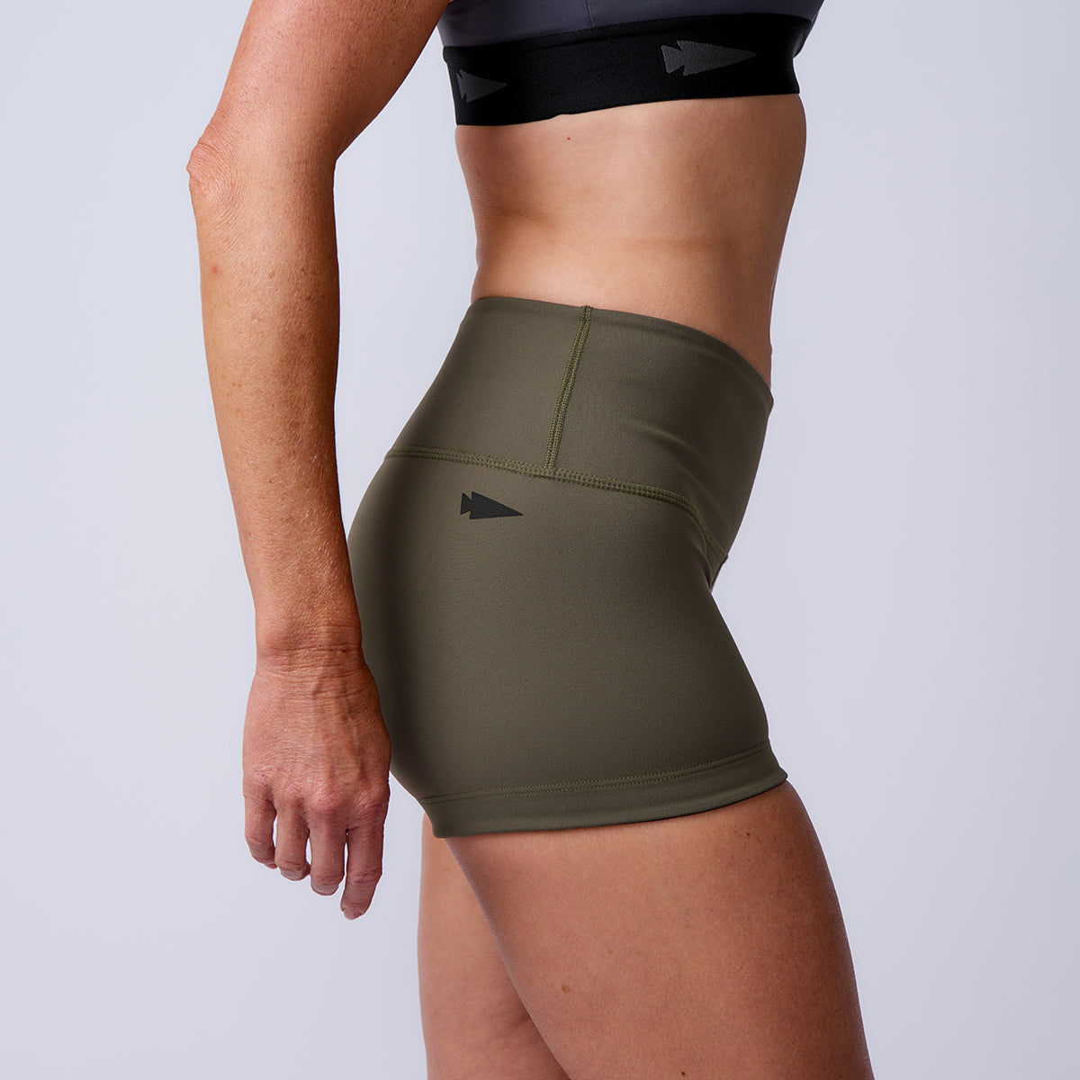 A woman wearing Women’s Squat Shorts - ToughFlex in high-waisted olive green and a black sports bra, shown from the side.