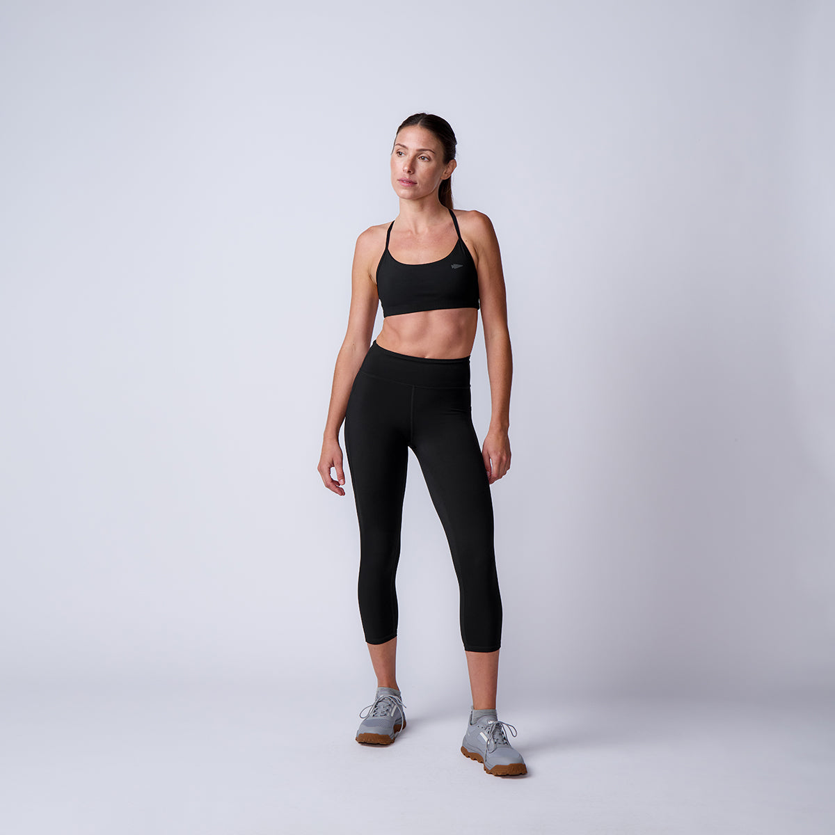 Woman in black Women’s Training Leggings Pocket - ToughFlex and sports bra stands against a plain light background, looking slightly to the side, showing off her durable leggings perfect for any workout.