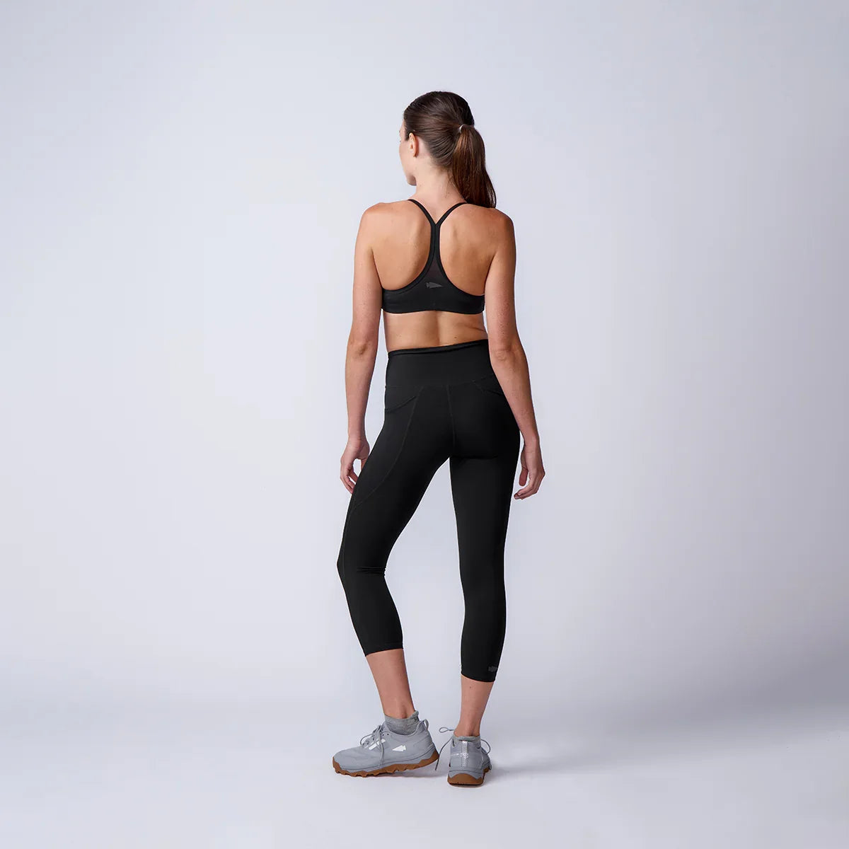 Woman wearing GORUCK black sports bra, leggings, and gray shoes, studio background