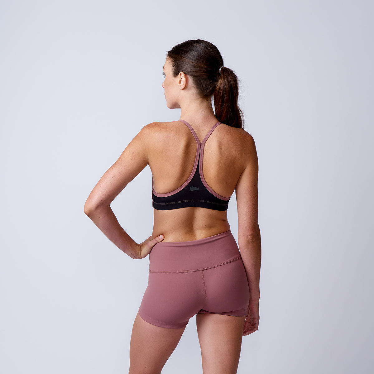 Woman in athletic wear stands with hand on hip, back facing camera against a plain light backdrop, highlighting high-waisted Women’s Squat Shorts - ToughFlex.
