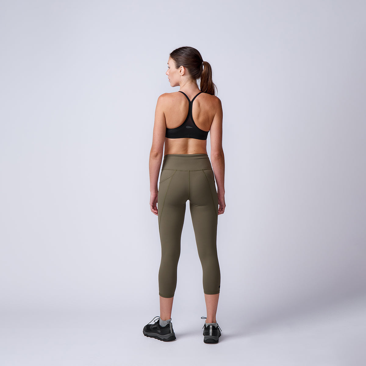 A woman in athletic wear stands facing away in a studio with a plain light background, modeling the high-waisted Women’s Training Leggings Pocket - ToughFlex.