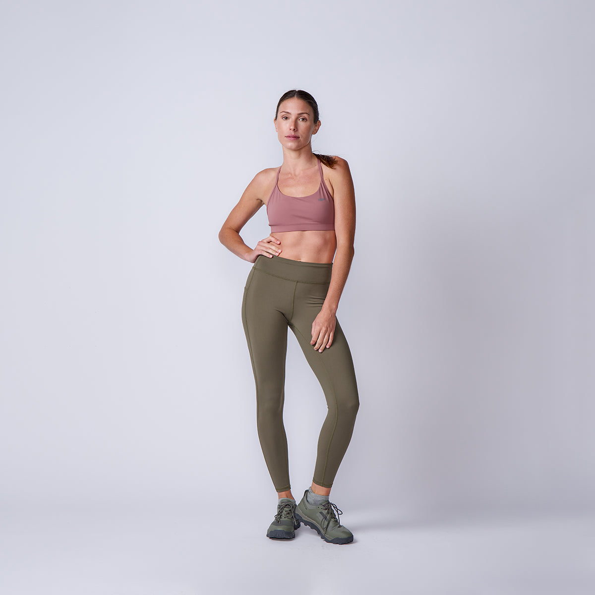 A woman in a mauve sports bra and olive green Women’s Training Leggings Pocket - ToughFlex stands against a plain light gray background.