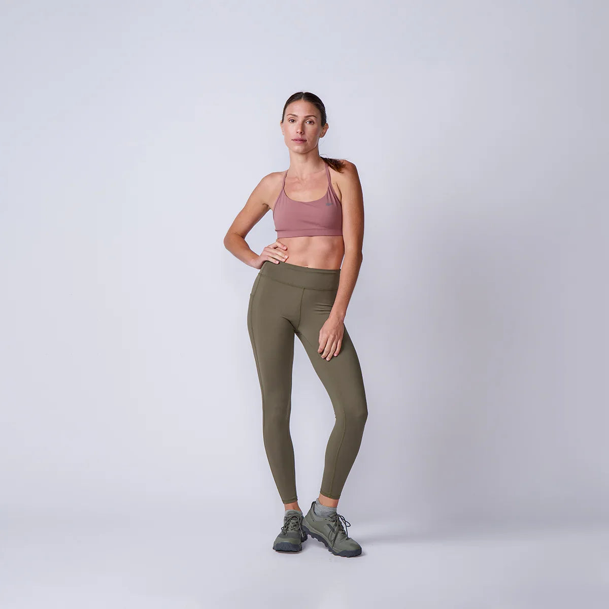 Woman wearing GORUCK olive green leggings, pink sports bra, and trail shoes in studio.