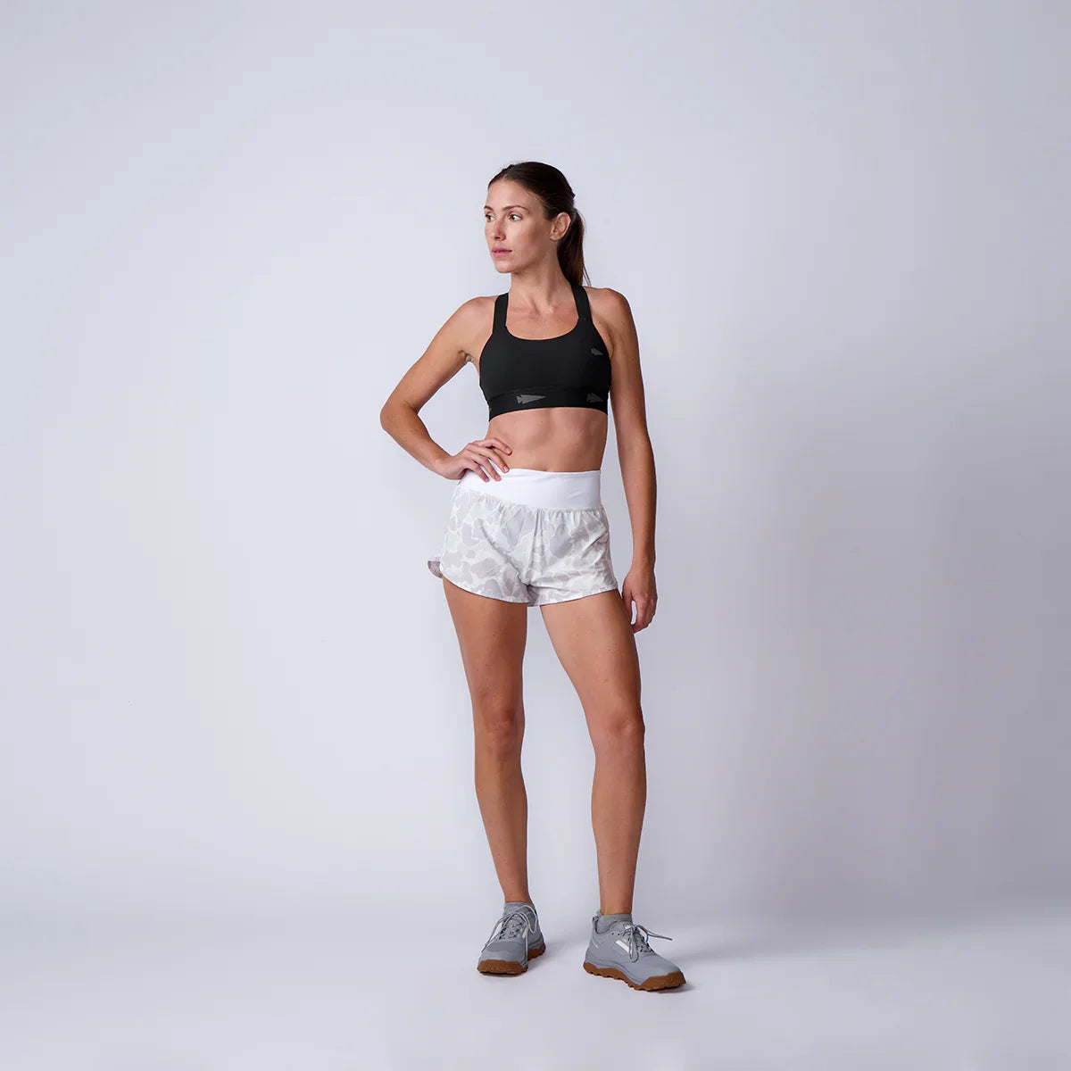 Athletic woman in black sports bra, white patterned shorts, and gray sneakers posing against light gray background