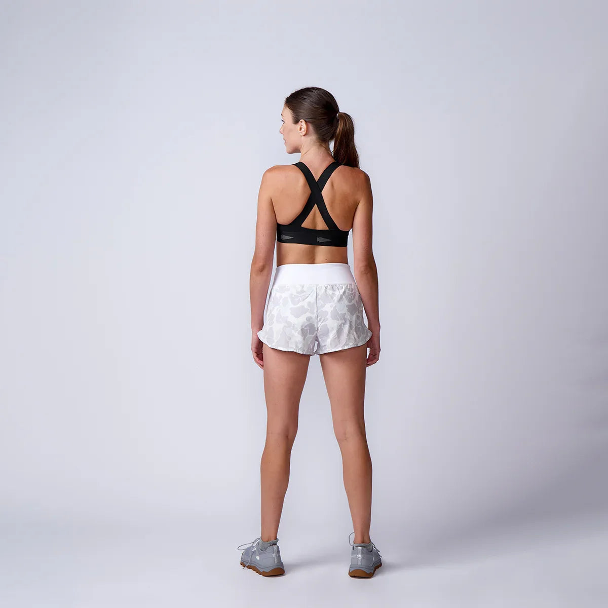 Woman in black sports bra and white camo print shorts standing facing away on grey background