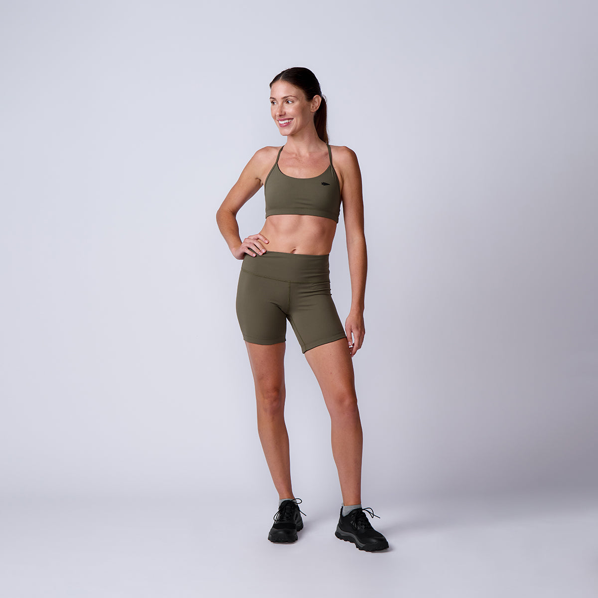 Smiling woman wears olive green Women’s Biker Shorts - ToughFlex and black sneakers, standing against a plain light gray background.
