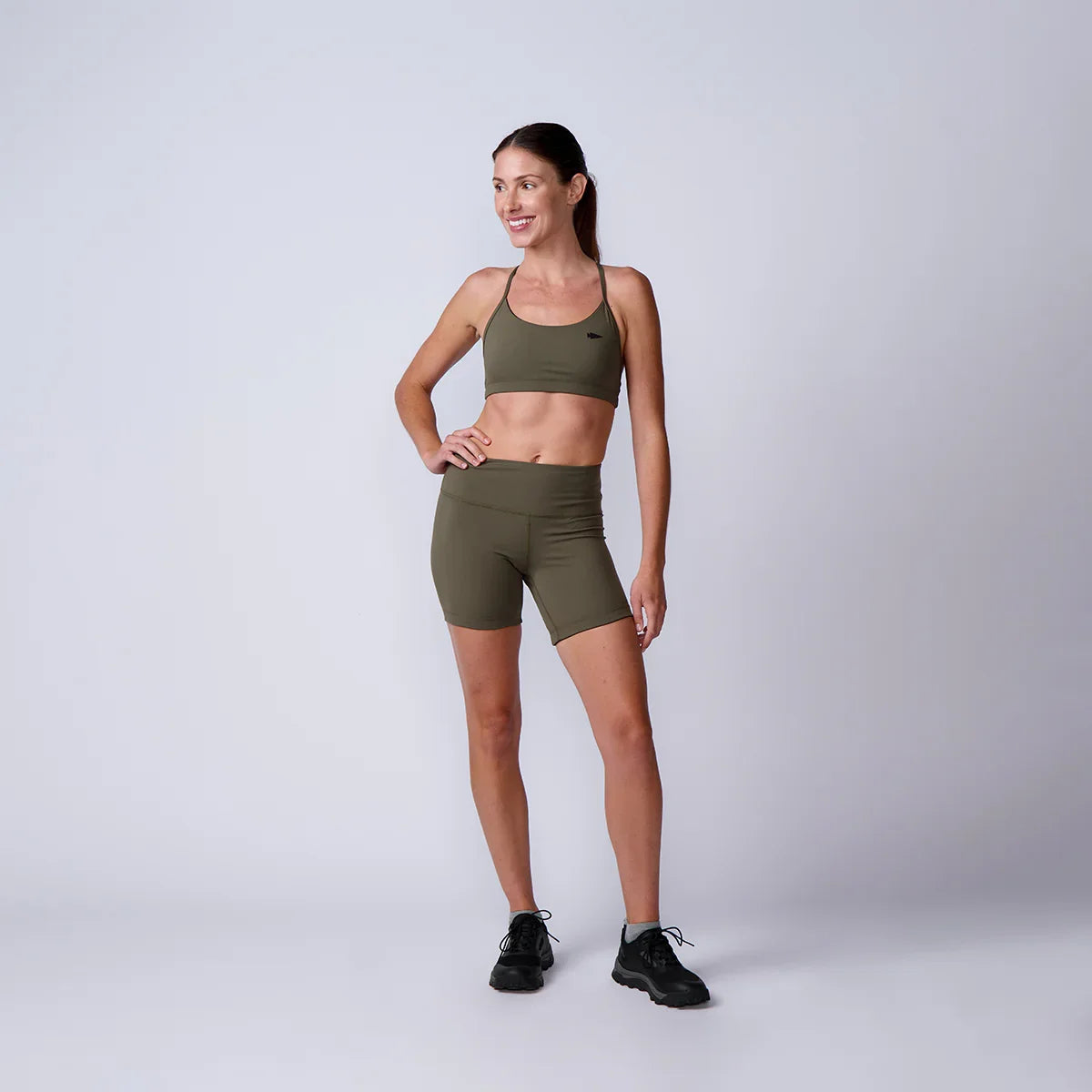 Smiling woman in olive green sports bra and shorts with black sneakers on plain gray background