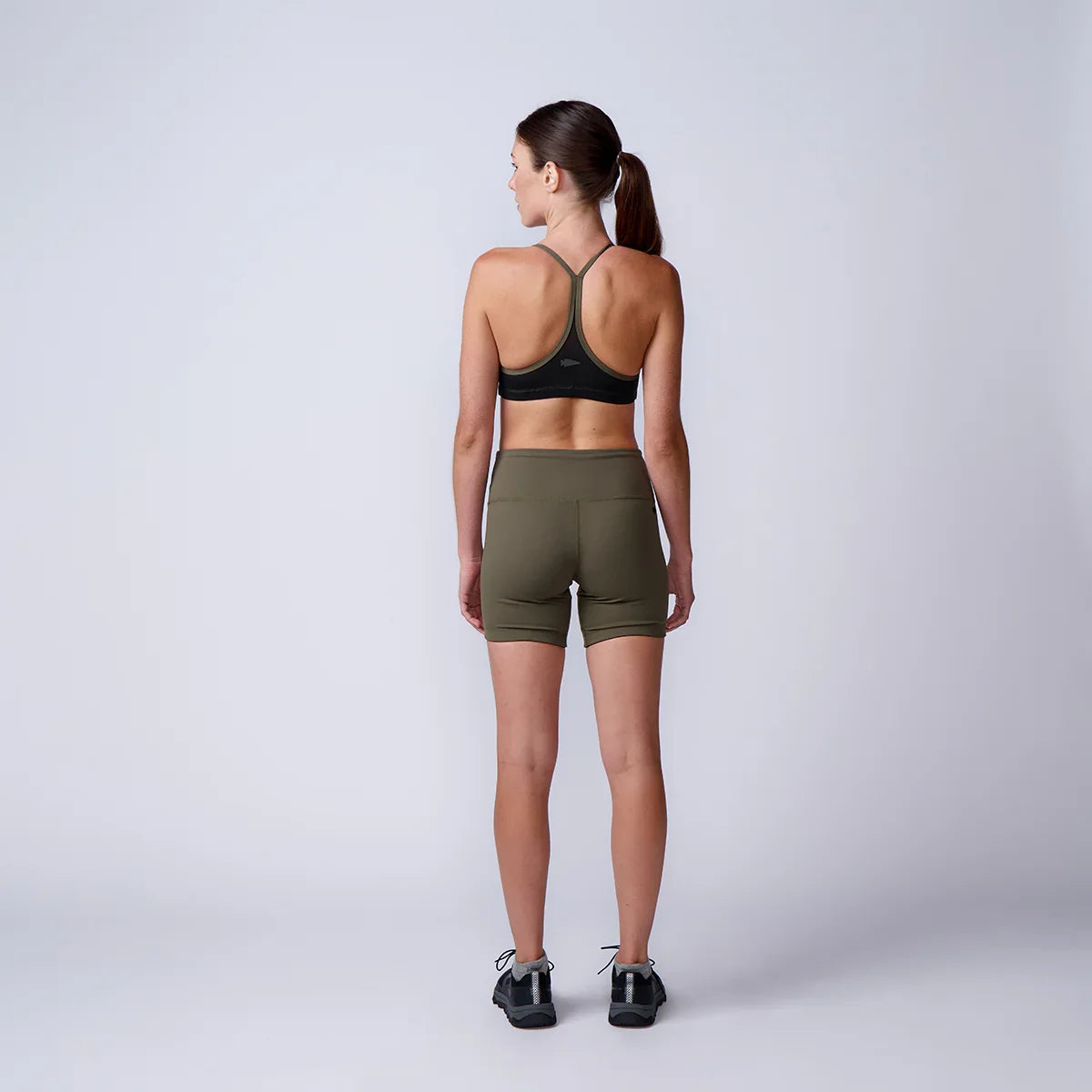 Athletic woman in olive green rucking shorts and black sports bra standing against gray studio background