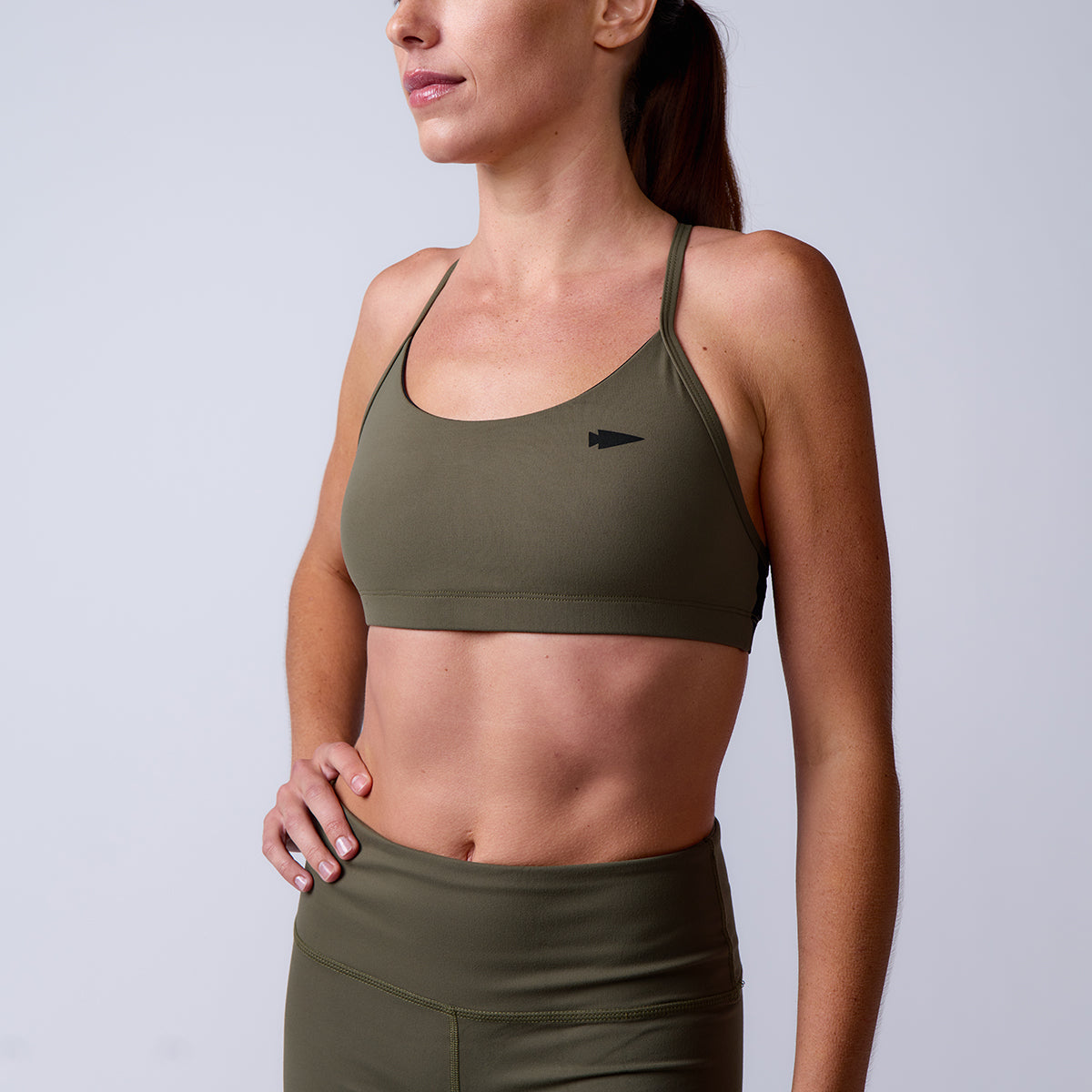 A woman wears the Stealth Bra - ToughFlex and matching leggings in olive green, featuring lightweight support, standing with one hand on her hip against a plain background.