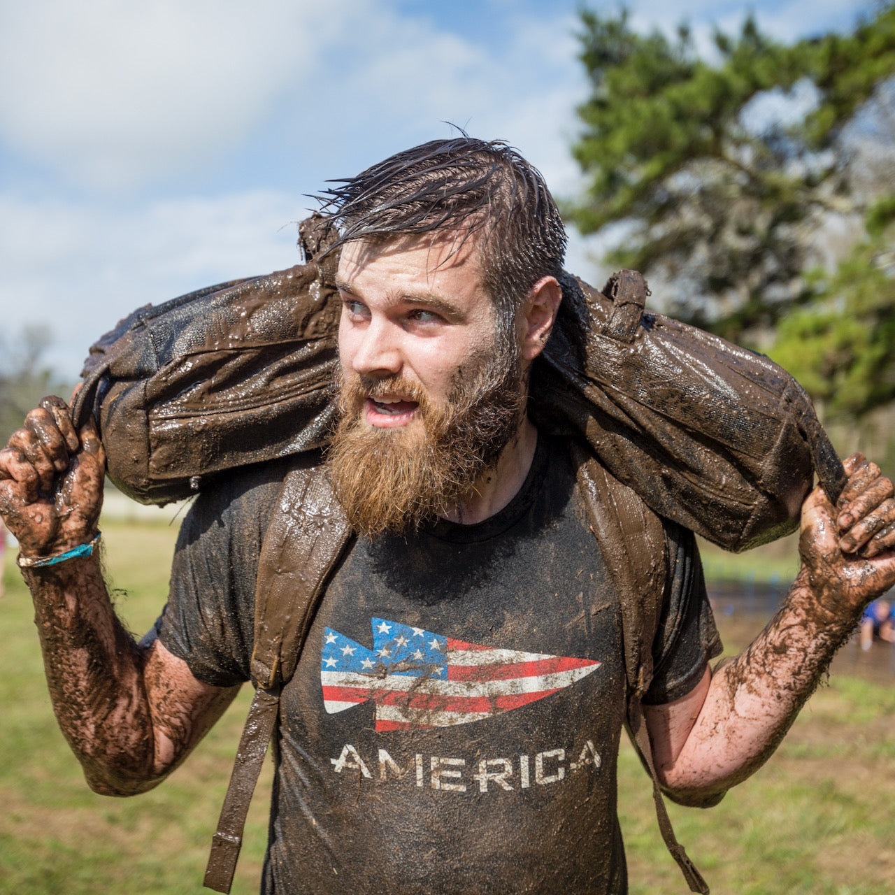 A bearded man with a muddy backpack wears the America Tee - Tri-BlendX outdoors, looking off to the side.