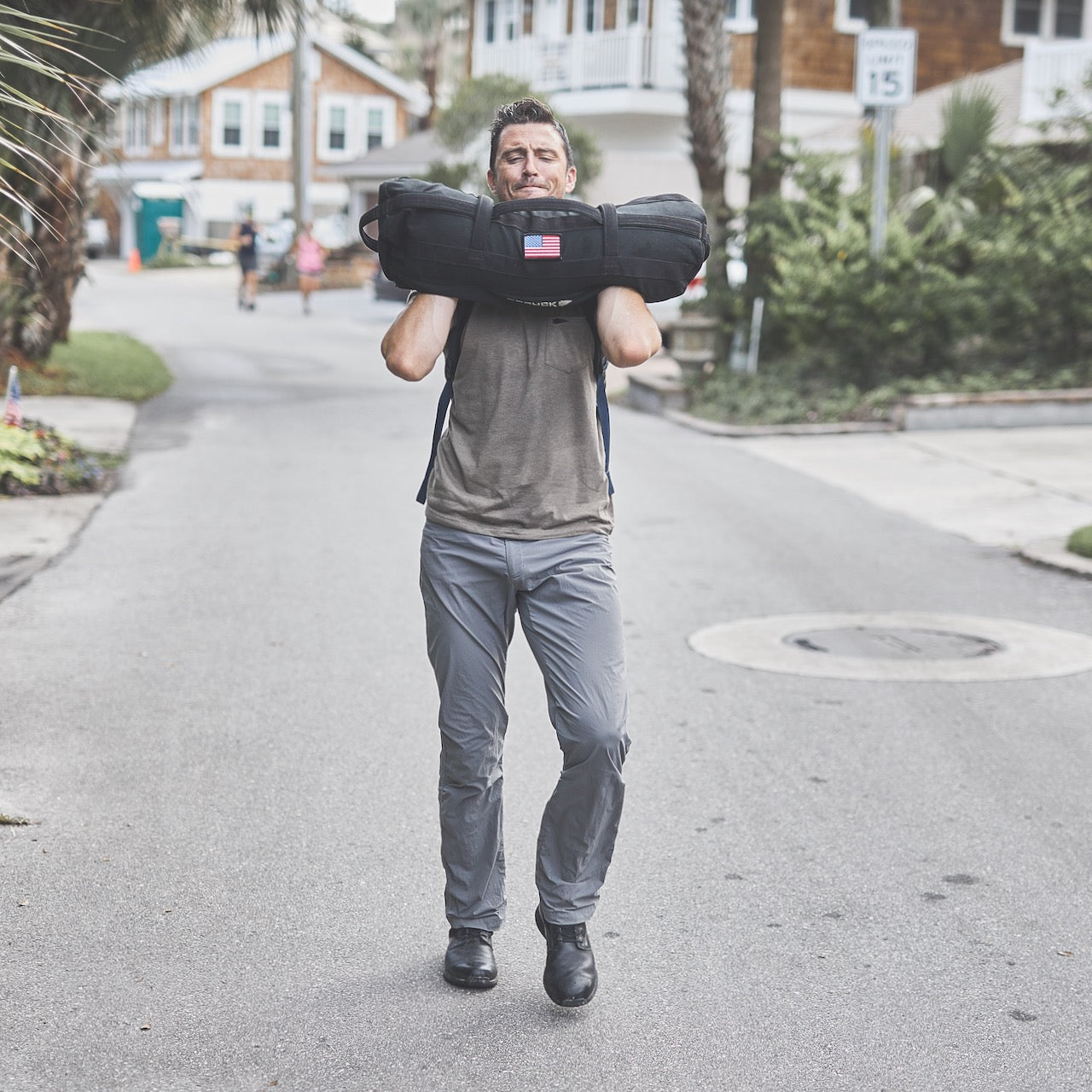 A man carries the Sandbag Kit (w/ Filler Bag) on his shoulders down a residential street, demonstrating its durability, strength, and lifetime guarantee for all your training needs.