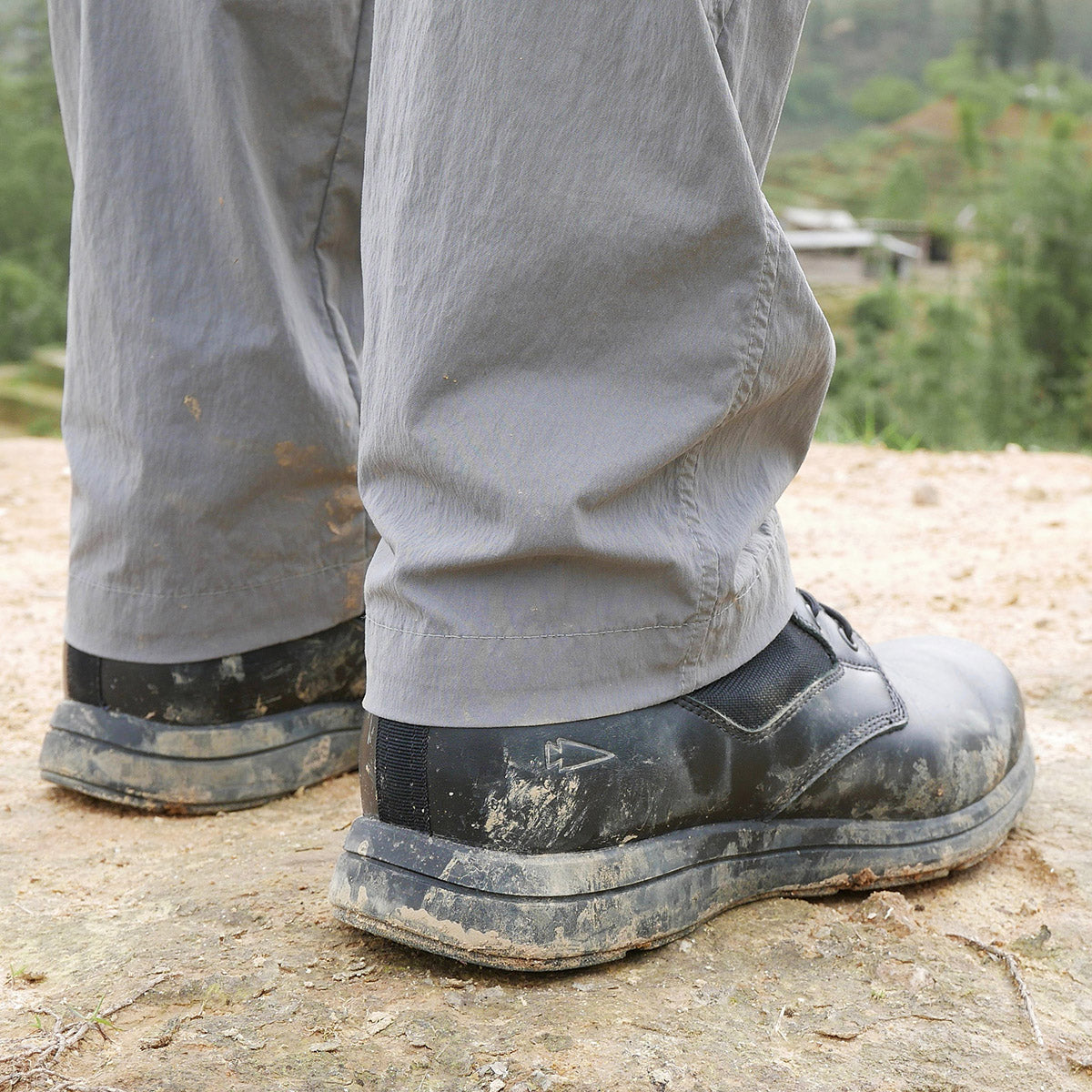 Person wearing gray pants and muddy black shoes standing outdoors on rocky ground.
