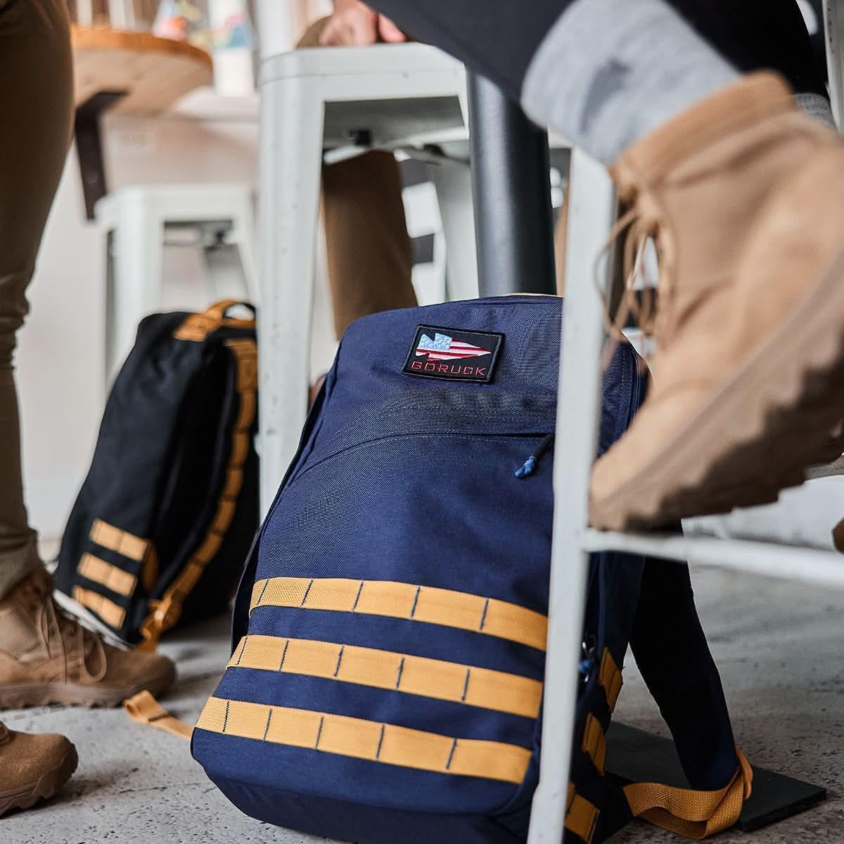 GORUCK navy blue rucksack with light gold straps under table, worn with tan boots, indoor setting