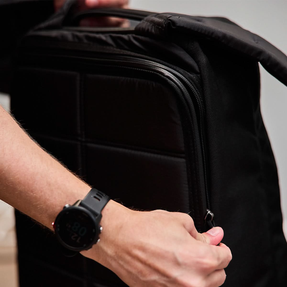A person wearing a black smartwatch is zipping up a black GR1 USA - Ballistic Nylon Cordura (The Original Ruck) 210D Back Panel, an everyday carry inspired by Special Forces.