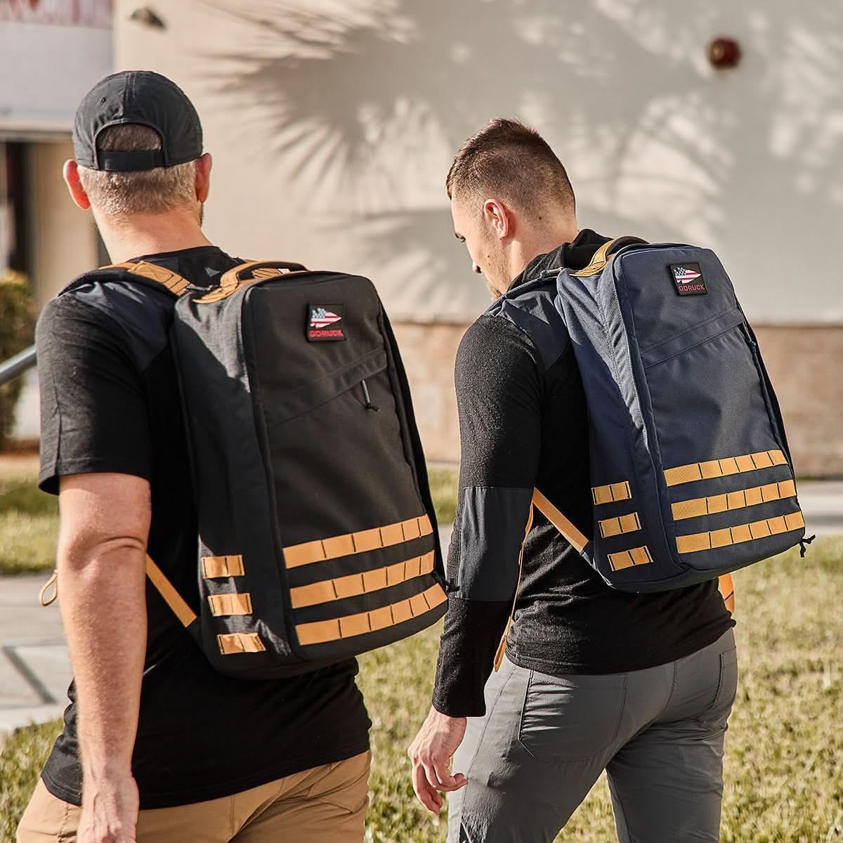 Two men wearing GORUCK rucksacks, one black and one navy, walking outdoors in casual activewear