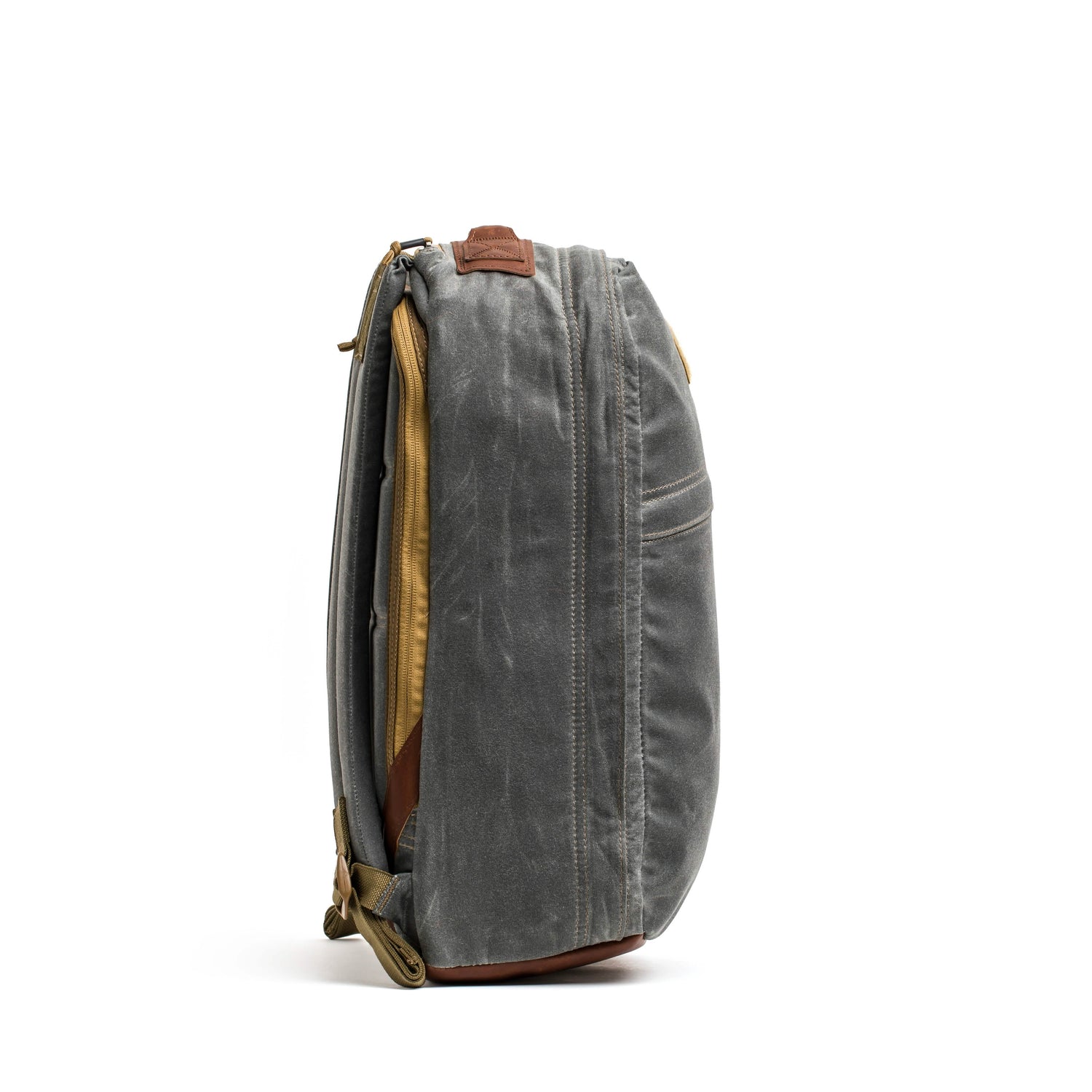 The GR1 USA - Heritage backpack by GORUCK, crafted from gray, waxed duck canvas with brown leather accents, stands upright against a white background. From the side view, you can see its zippered main compartment and adjustable shoulder straps. The heritage rucksack boasts a vintage look with a slightly worn texture.