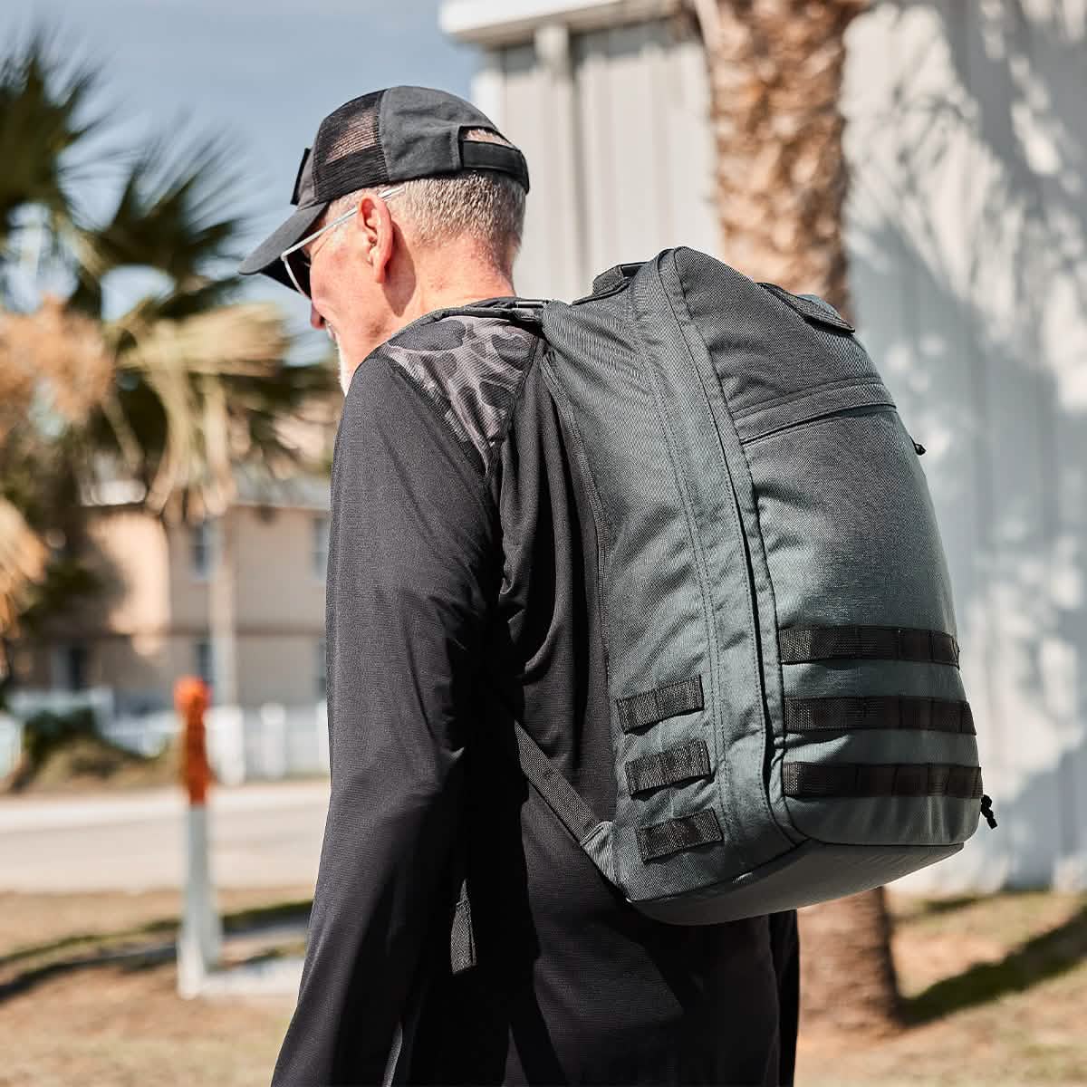 A person in a black cap and shirt, carrying the large gray GR1 USA - Cordura backpack, originally designed by GORUCK and often preferred by Special Forces, walks outdoors. The scene is set against a backdrop of palm trees, a building, and a clear sky.