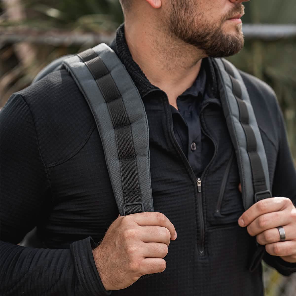 A man wearing a black long-sleeve shirt is holding the straps of the GR1 USA - Cordura, also known as The Original Ruck by GORUCK, celebrated for its rugged durability in the GORUCK Challenge. The backpack features gray and black padded straps. In the background, green foliage blurs softly.