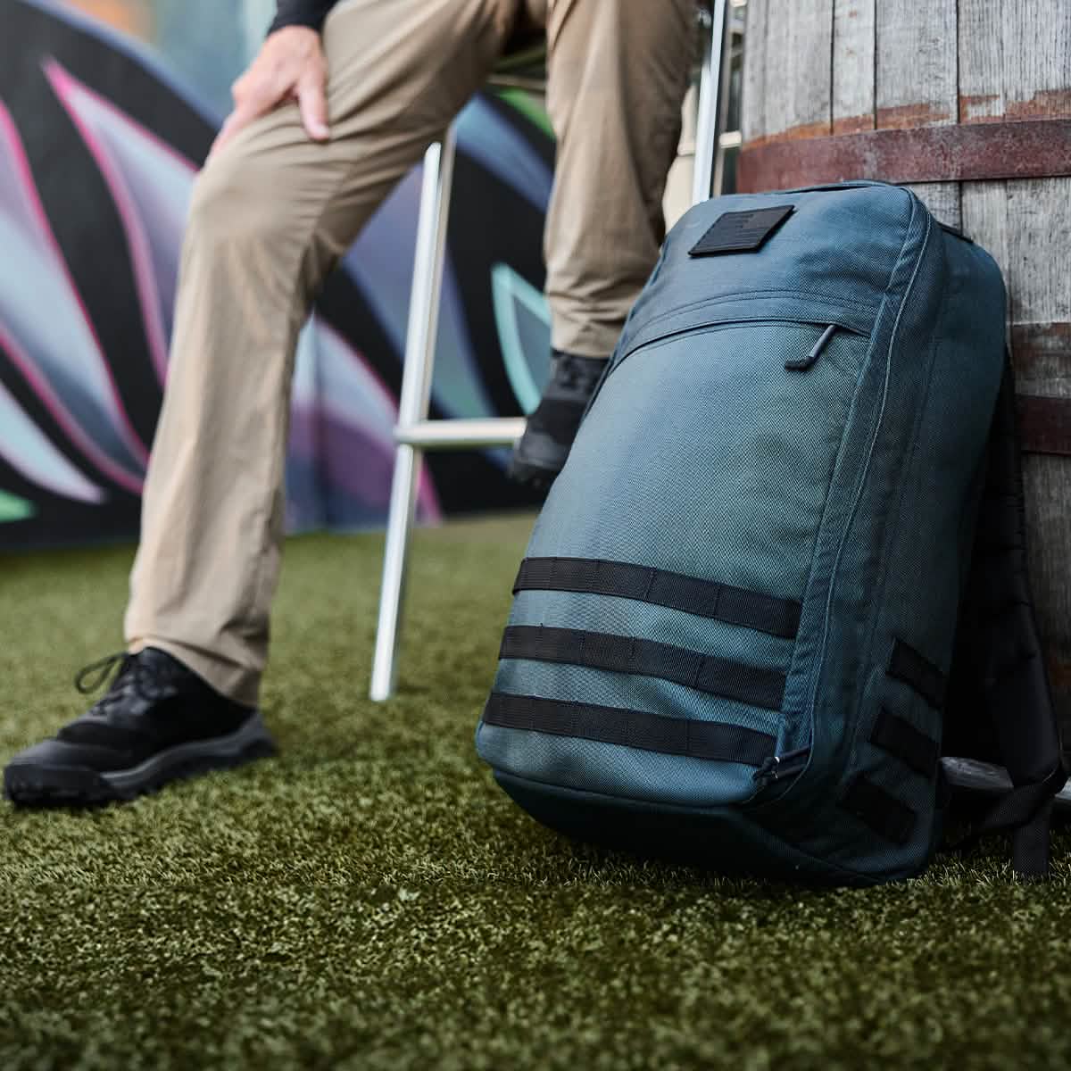 A person sits on a stool on artificial grass, clad in khaki pants and black shoes that evoke the rugged style of a Special Forces operative. Beside them rests a GR1 USA - Cordura (The Original Ruck) backpack by GORUCK, featuring distinctive black horizontal straps as it leans against a barrel. In the background, vibrant graffiti adds a splash of color.